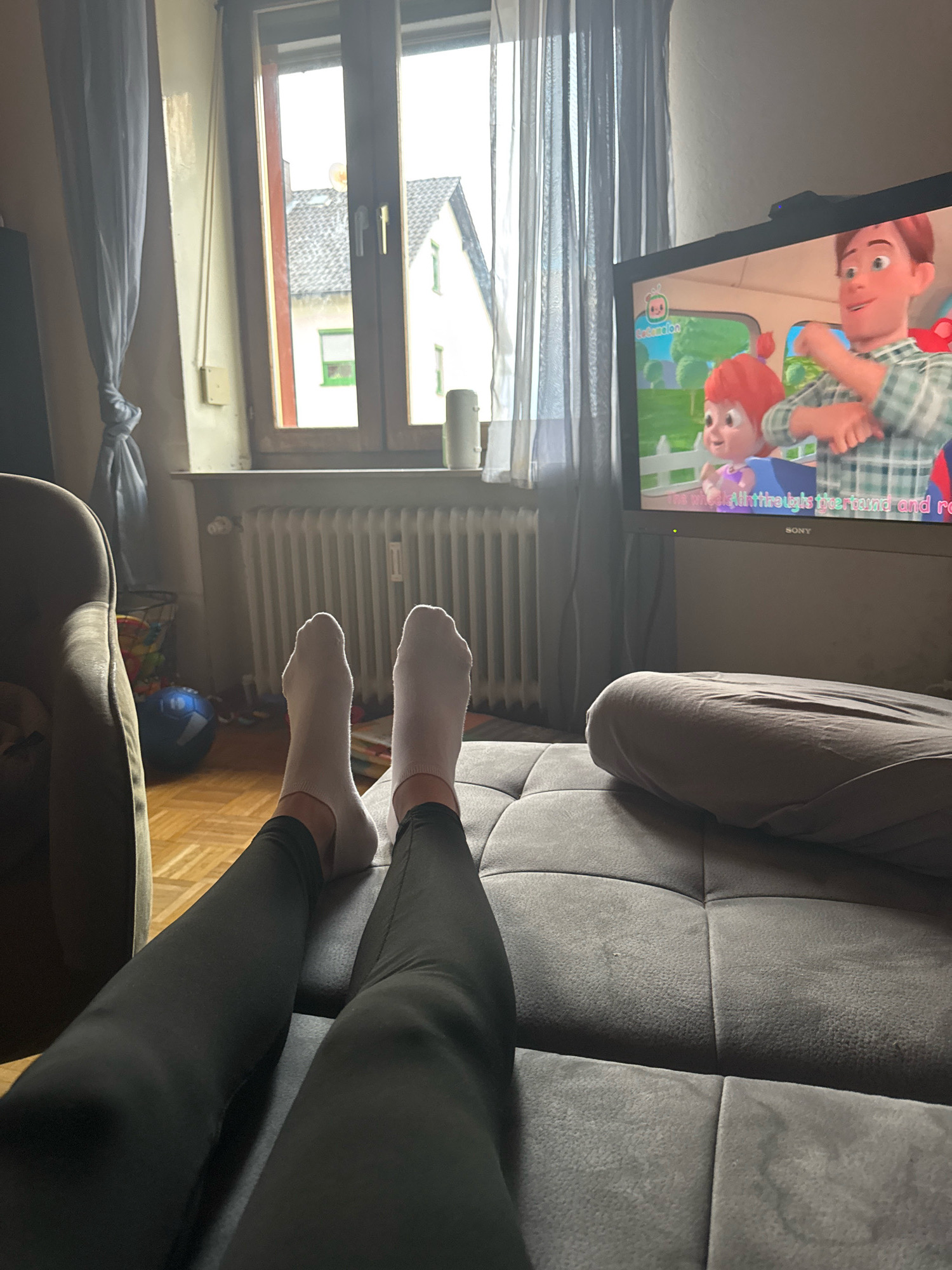 The image depicts a living room scene in Überherrn, Germany. A person is lying on a dark grey couch, with their legs and feet, clad in white socks and black leggings, extended towards the camera. In the background, a television screen displays a cartoon from the show "CoComelon." To the left of the television, sheer grey curtains hang by a window, revealing houses and a cloudy sky outside. A radiator is visible below the window, and a basket containing children's toys, including a blue ball, sits on a wooden floor. A plush armchair is partially visible on the left side of the frame. The overall atmosphere is relaxed and domestic.