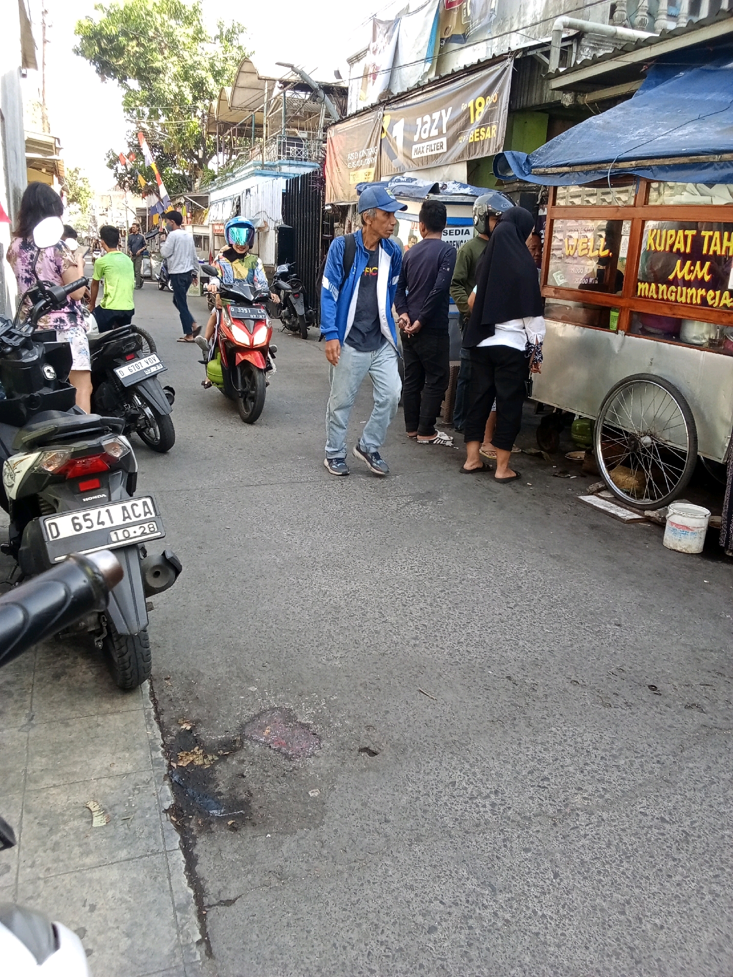 A street scene in Bandung City, Indonesia, features several individuals and vehicles. A man in a blue jacket and blue cap walks on the asphalt road, his hands clasped behind his back. To his left, a person in a black hijab and white shirt stands near a food cart. Behind them, a group of people are visible, including a person in a green t-shirt. Several motorcycles are parked on the left side of the street. The food cart has signage that appears to be in Indonesian. The road surface is asphalt, and there are some puddles and debris. The environment suggests an outdoor market or a busy street in an urban area.