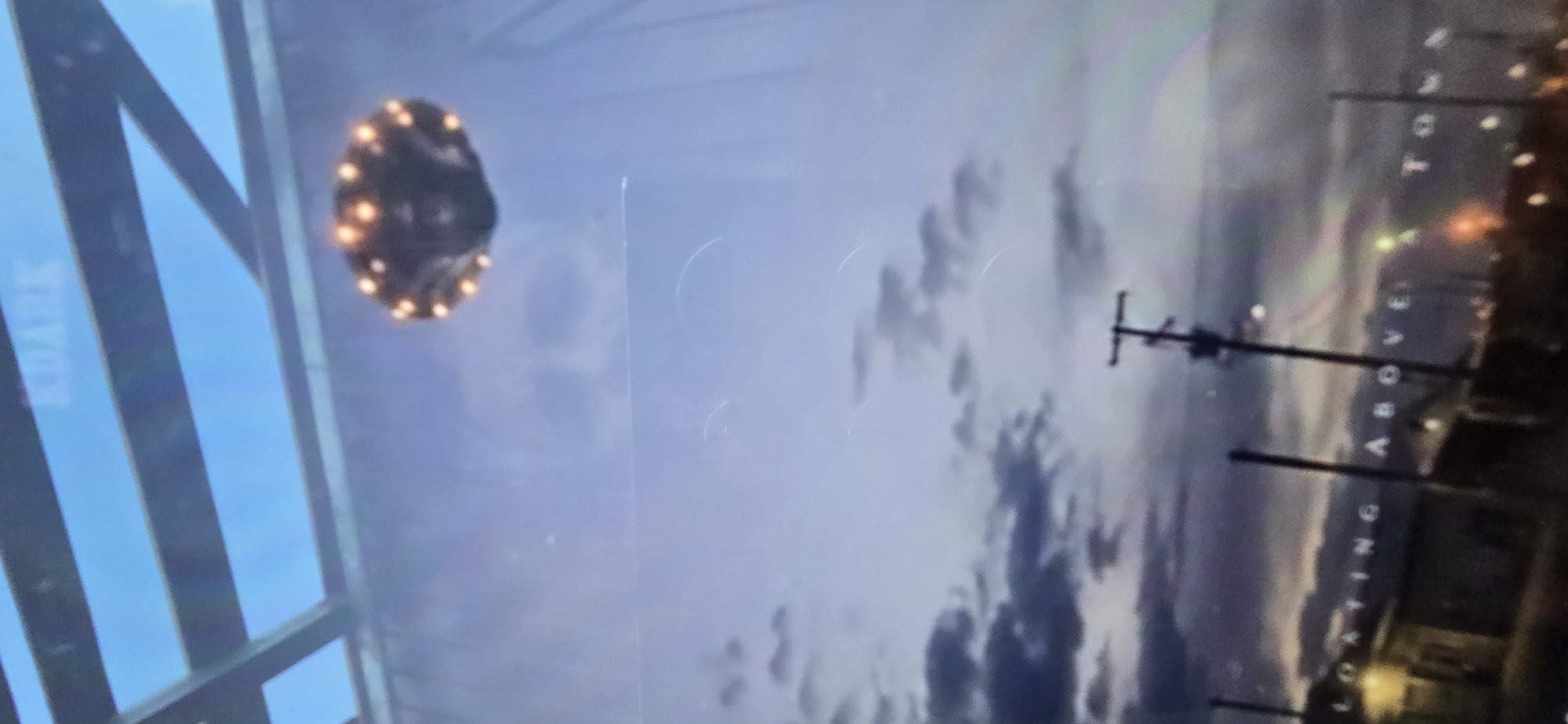This media file depicts a hazy, atmospheric scene, possibly a screen capture from a video. The central focus is an unidentified flying object, dark and oval-shaped, with a series of bright orange lights encircling its perimeter. It appears to be hovering in a sky filled with diffused light, suggesting either dawn, dusk, or overcast conditions.
To the left of the UFO, a portion of a structure with dark vertical beams and a blue, possibly reflective surface is visible, with the word "KURGAN" vertically oriented on a white stripe. This suggests a connection to the city of Kurgan, Russia.
To the right of the UFO, the scene transitions to what appears to be a landscape with a utility pole and wires. In the background, lights from a town or city are dimly visible. The sky is a mix of grey and purple hues, with swirling cloud formations that create a somewhat dramatic or ethereal effect. White text is superimposed vertically, reading "LOSTING ABOVE TOWN".
The overall impression is one of mystery and perhaps unease, with the glowing UFO and the subdued lighting contributing to a sense of the unknown. The weather appears to be hazy or foggy, obscuring finer details of the background. The time of day is ambiguous due to the lighting conditions, but the presence of artificial lights in the background suggests it is not broad daylight.