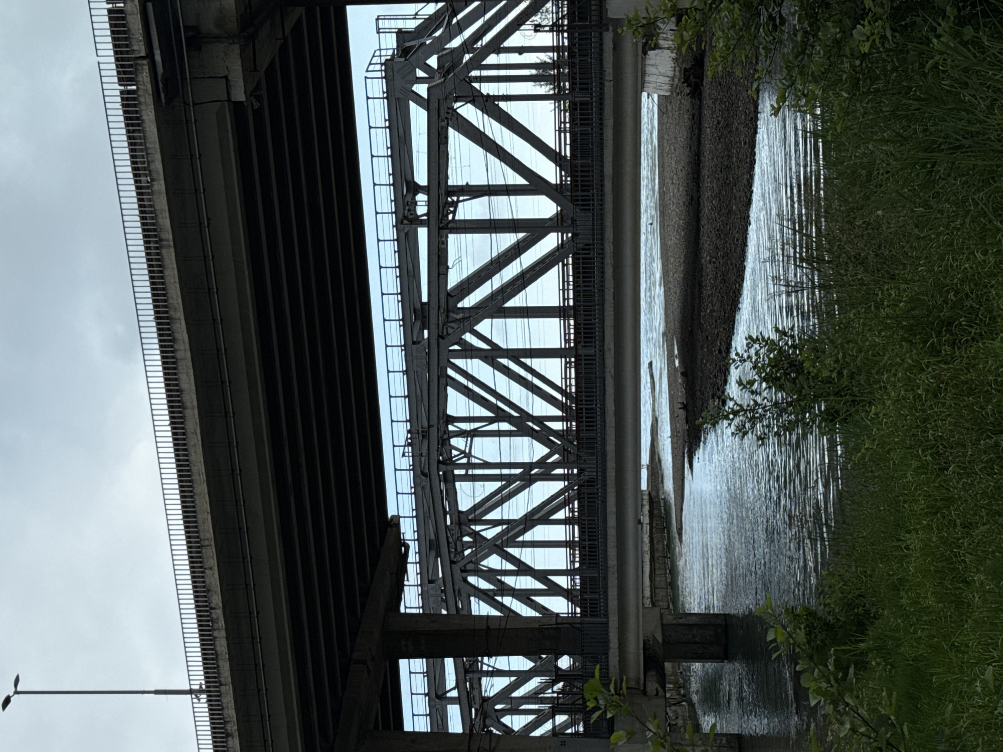 The image shows two bridges over a body of water. One bridge is a girder bridge with a regular pattern of vertical supports beneath the deck, and a walkway with a railing. The other bridge appears to be a truss bridge with a complex web of steel beams. There is vegetation on the right side of the image, suggesting the edge of a riverbank or shoreline. The water is reflective, indicating a relatively calm surface. A light pole is also visible, further indicating the presence of infrastructure. The location appears to be urban.