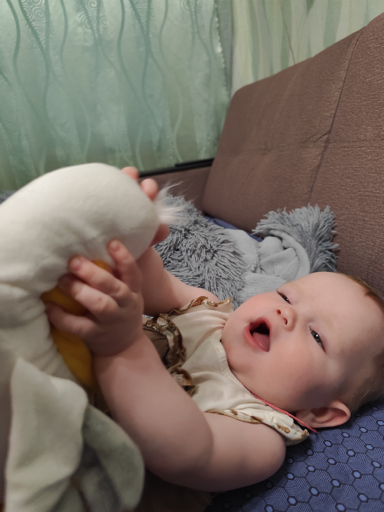 A baby is lying on a blue patterned surface, possibly a bed or a sofa, holding a white and yellow stuffed animal. A gray, fluffy blanket or pillow rests behind the baby. The background includes a brown couch and a green patterned curtain. The location of the scene is indiscernible and could not be identified as Фоки, Russia.