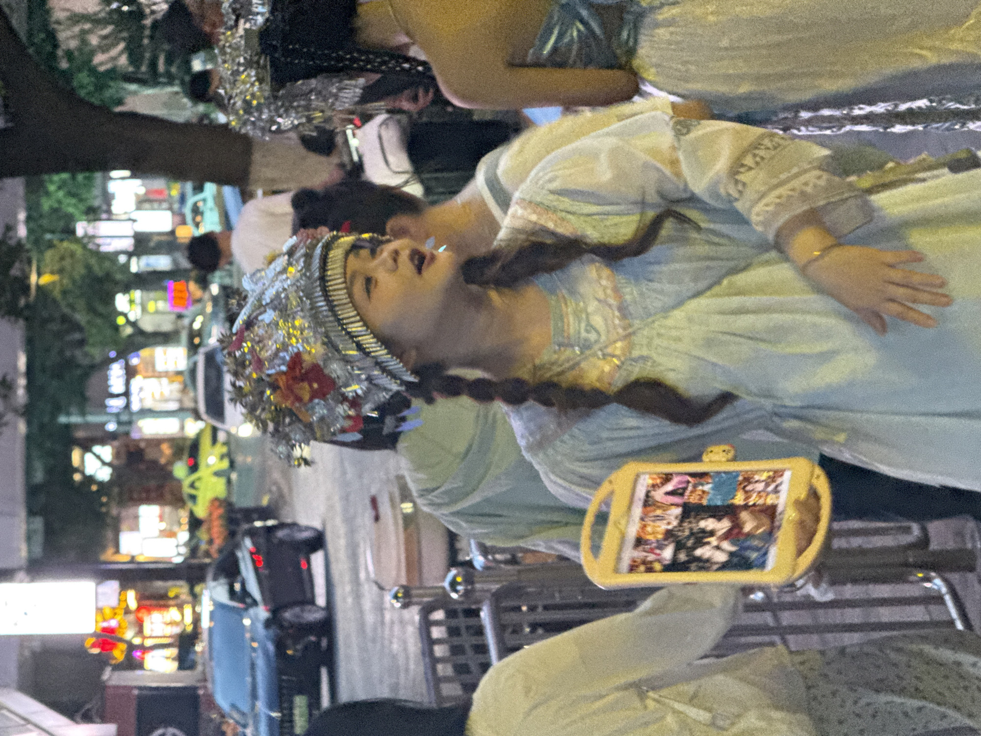 The image shows a group of people in what appears to be a city street at dusk or night, likely in Yuzhong District, China, given the context provided. The foreground features a young woman wearing an ornate, silver, and red headdress and a light blue traditional-looking dress with intricate embroidery on the sleeves. She is holding a yellow, rectangular object, possibly a tablet or phone, displaying several images of people, some of whom appear to be dressed in similar costumes. Her mouth is slightly open as if she is speaking or singing.
Behind her, other individuals are visible, also seemingly adorned in festive or theatrical attire, with one person in the mid-ground wearing a shimmering silver garment. In the background, a busy street scene unfolds with cars, including a black sedan and a bright yellow car, suggesting urban traffic. Buildings with illuminated signs and lights are visible, indicative of a commercial or entertainment district. The overall atmosphere seems to be that of a public gathering or performance, possibly a street festival or parade.