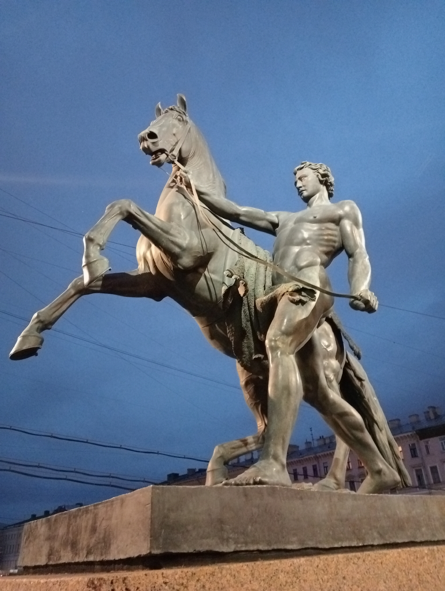A bronze equestrian statue of a man taming a horse stands on a stone pedestal. The man is depicted shirtless and muscular, with a determined expression, holding the horse's bridle. The horse rears on its hind legs, appearing agitated. The sculpture is set against a deep blue, twilight sky. In the background, the outlines of buildings and utility wires are visible. The statue is likely "The Bronze Horseman," a monument to Peter the Great located on Senate Square in Saint Petersburg, Russia.
