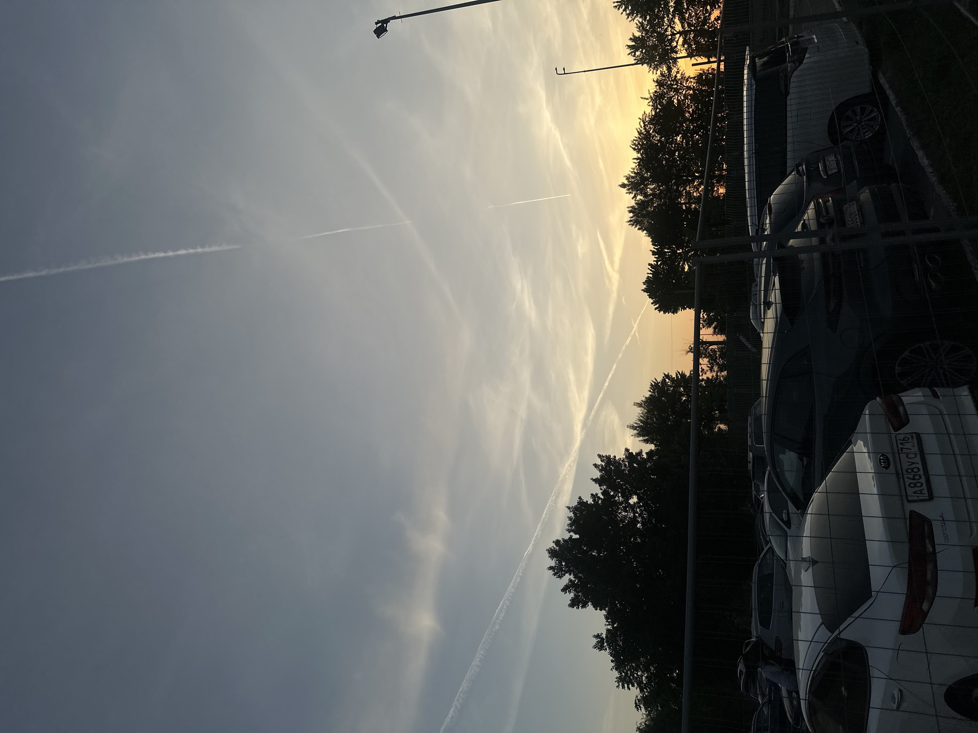The image depicts a sky with vapor trails crisscrossing above a car park in Kazan, Russia. The sky fades from grey to yellow near the horizon, suggesting a sunset or sunrise. Along the right edge of the frame, silhouetted trees and streetlights are visible, lining the area next to a wire fence which separates the car park from the trees. Several cars are parked behind the fence, and a Russian license plate "A868YC716" is visible on a white car in the foreground. No people are clearly visible.
