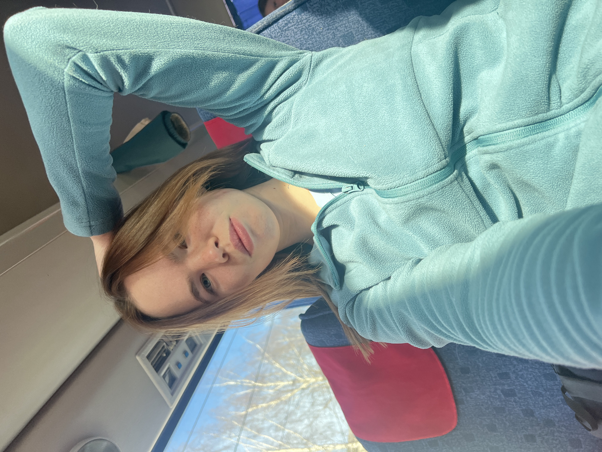 A woman with light brown hair is reclining on a seat. She is wearing a light blue fleece jacket. She is looking at the camera with a neutral expression. She appears to be on a bus or train with seats upholstered in blue and red. In the background, outside the window, are bare trees. The city is identified as Berezniki, Russia.