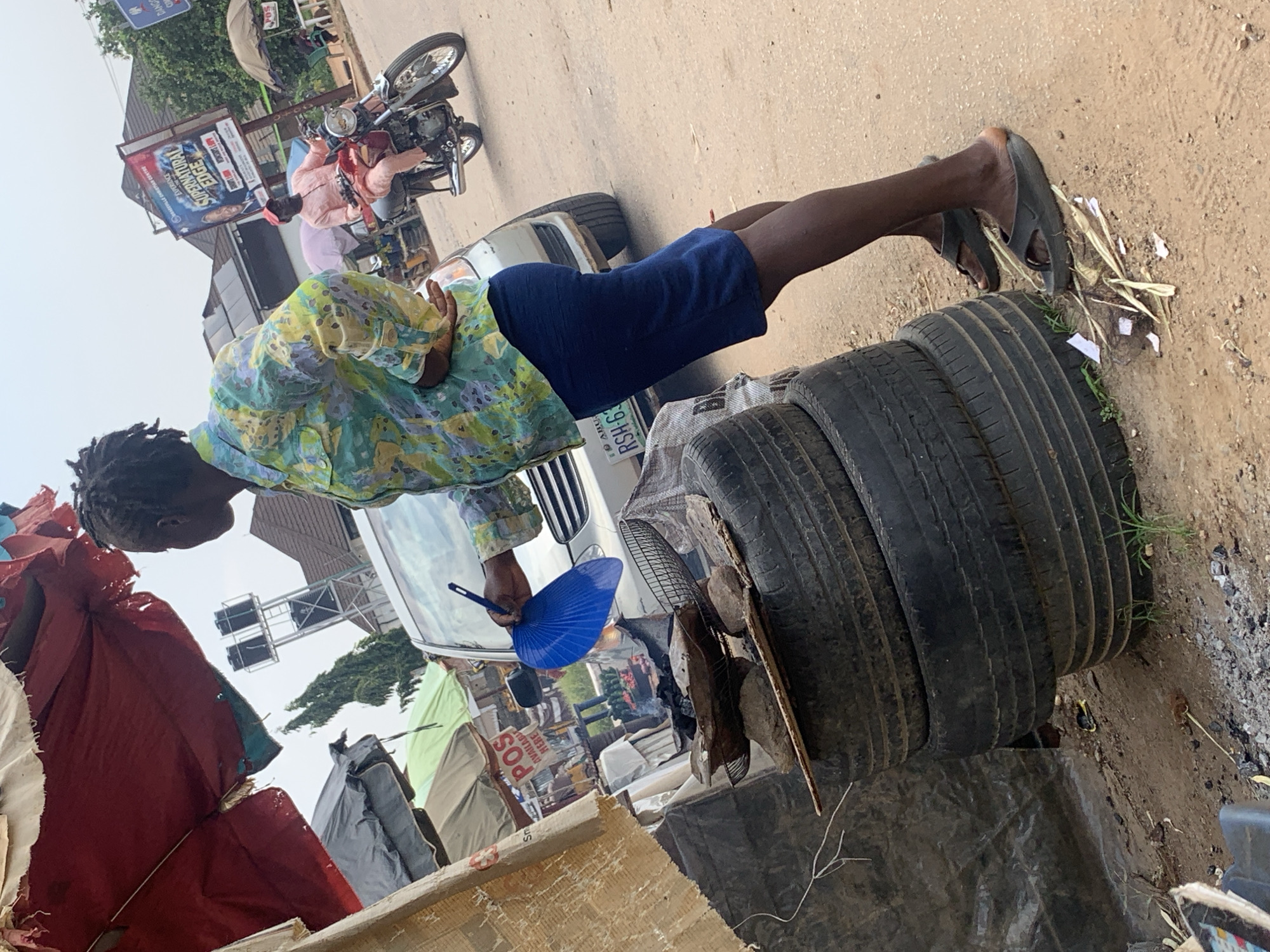 This media file depicts a street scene in Abuja, Nigeria, likely during the daytime given the bright lighting. The primary subjects are a young man with dark, braided hair, wearing a vibrantly patterned short-sleeved shirt and blue shorts, and a stack of used tires. He is leaning on the tires, holding a blue hand fan, possibly to cool himself. His posture suggests a moment of rest or waiting.
In the background, various elements suggest a busy urban environment. A motorcycle with a rider wearing a pink shirt is visible, as well as a white vehicle. Signs with text are present, including "POS AVAILABLE HERE" and a sign for "EXPERIENCE SUPERNATURAL EDGE," indicating commercial activity. Tents or makeshift shelters are also scattered around, hinting at a market or informal settlement area. The ground is dusty and appears to be a dirt road or lot, typical of some areas in Abuja.
The overall impression is one of everyday life in a bustling Nigerian city, with elements of commerce, transportation, and informal living spaces coexisting. The weather appears to be warm, as suggested by the man's attire and the hand fan. There are no strong emotional cues evident, but the scene conveys a sense of typical daily activity.