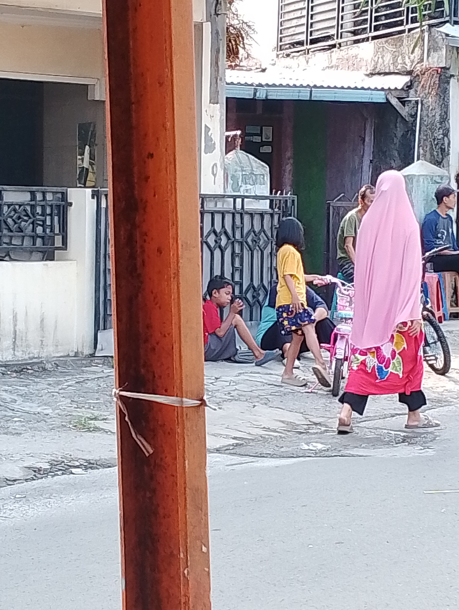 A group of people are gathered on a street in Bogor, Indonesia. In the foreground, a wooden pole with a white tie around it stands prominently. To the right of the pole, a person wearing a pink hijab and a brightly colored skirt walks away from the camera. A small pink bicycle is positioned near them. Further to the left, a child in a yellow shirt and patterned shorts stands near the bicycle. To the left of the child, a person in a blue shirt is sitting down. In the background, a boy in a red shirt and grey pants is sitting on the ground, looking at his phone. Behind him, a man in a green shirt is visible. The environment appears to be a street with a sidewalk and buildings in the background. The lighting suggests it is daytime, with sunlight casting shadows.