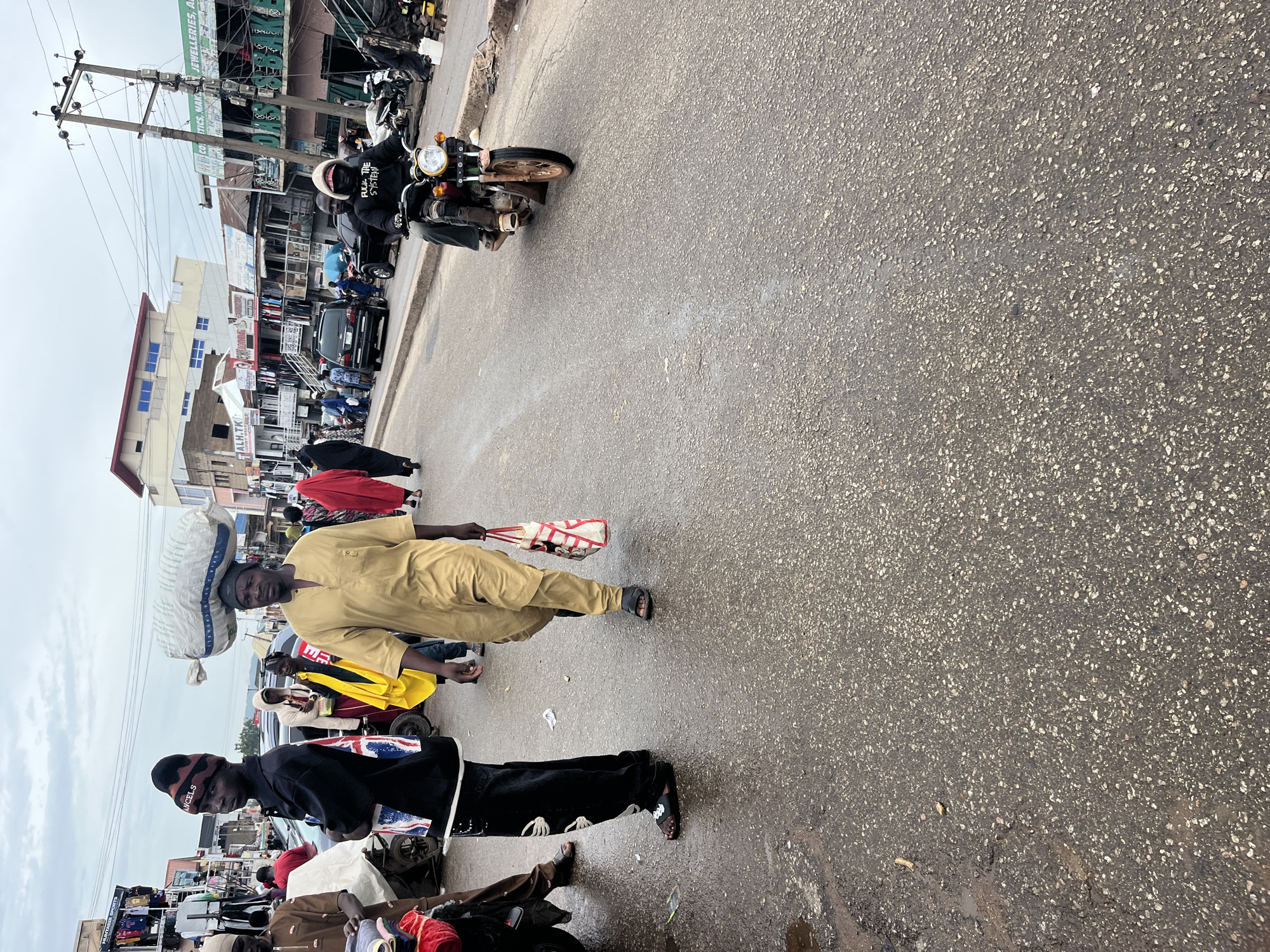 A street scene in Jos, Nigeria, features a road wet from recent rain. In the foreground, a man wearing a tan tunic and pants walks away from the camera, carrying a red and white bag. To his left, another man in a black long-sleeved shirt and black pants with white accents, wearing a black beanie, walks towards the camera. He has a red and white bag over his shoulder. Behind them, a motorcyclist in a black hoodie rides away from the camera on a brown motorcycle. Numerous people are visible in the background, some walking and others standing near shops. Buildings line the street, with a prominent building featuring a blue-capped roof and several stories. Overhead power lines and poles are also present. The sky is overcast with clouds.