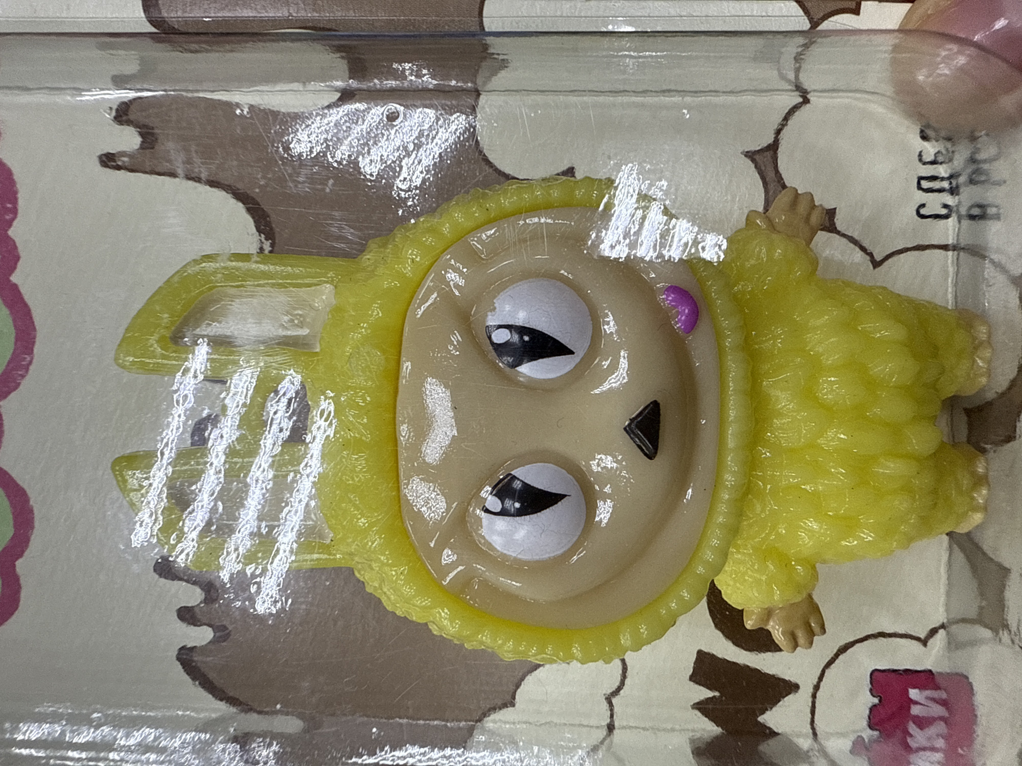 The image shows a yellow, furry plastic toy in a clear plastic package. The toy has a round face with large, white eyes with black pupils, a triangular black nose, and what appears to be a purple dot on its forehead. The background of the packaging has a pattern of cartoonish shapes, some of which are brown. The toy is in a sealed package, likely for retail sale. Some text in Russian is partially visible in the lower right corner and top right corner of the image. Based on the presence of Russian text, the item could be located in Votkinsk, Russia.