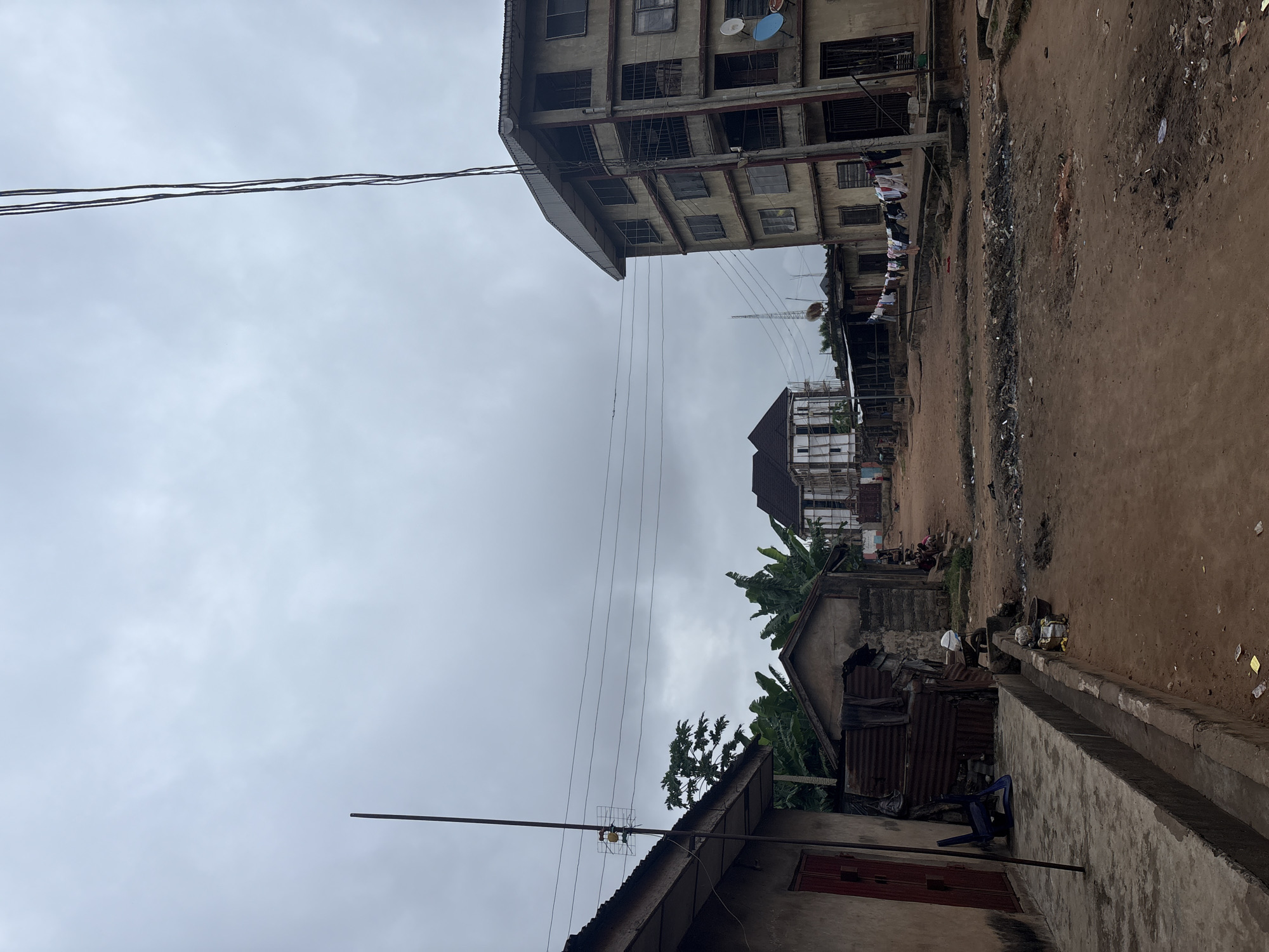 The image depicts an outdoor scene in Onitsha, Nigeria, under an overcast sky suggesting it might be a cloudy day or late afternoon. The foreground is dominated by a rough, dirt path bordering a concrete structure. Several buildings are visible, including a multi-story concrete structure with numerous windows and balconies, some of which appear broken or unfinished. Clothes are hanging on a line strung between two buildings, suggesting a residential area. Another building, with a corrugated metal roof, is partially visible, and a blue plastic chair sits on the concrete. Further back, more buildings and vegetation, including banana trees, are present. The ground is littered with debris and trash, indicating a less-than-pristine environment. There are no clear indications of specific activities or events taking place, and no visible text. The overall impression is one of a dense urban environment with signs of everyday life, but also neglect and poverty.