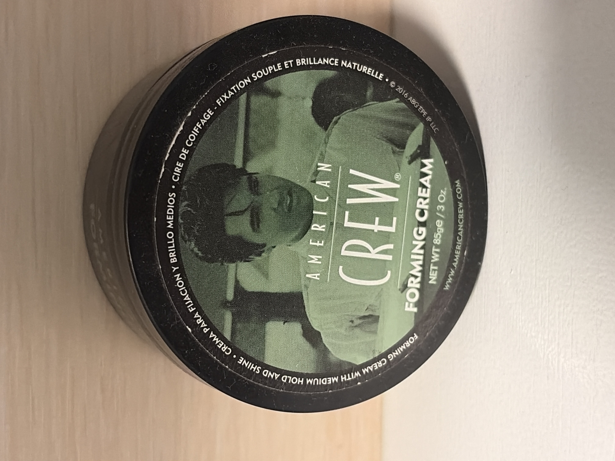 The image shows a black container of American Crew Forming Cream on a light-colored surface. The container is round and cylindrical, with the product label visible on the top. The label features a black and white image of a man, the words "AMERICAN CREW" in a large, stylized font, and the words "FORMING CREAM" below it. Additional text circles the perimeter of the lid, including "CREMA PARA FIJACIÓN Y BRILLO," "CIRE DE COIFFAGE - FIXATION SOUPLE ET BRILLANCE NATURELLE," "FORMING CREAM WITH MEDIUM HOLD AND SHINE," the net weight (85ge/3 oz.), and the website "WWW.AMERICANCREW.COM." The copyright date "© 2016 ABG EPE IP LLC" is also printed on the lid. The scene appears to be indoors.