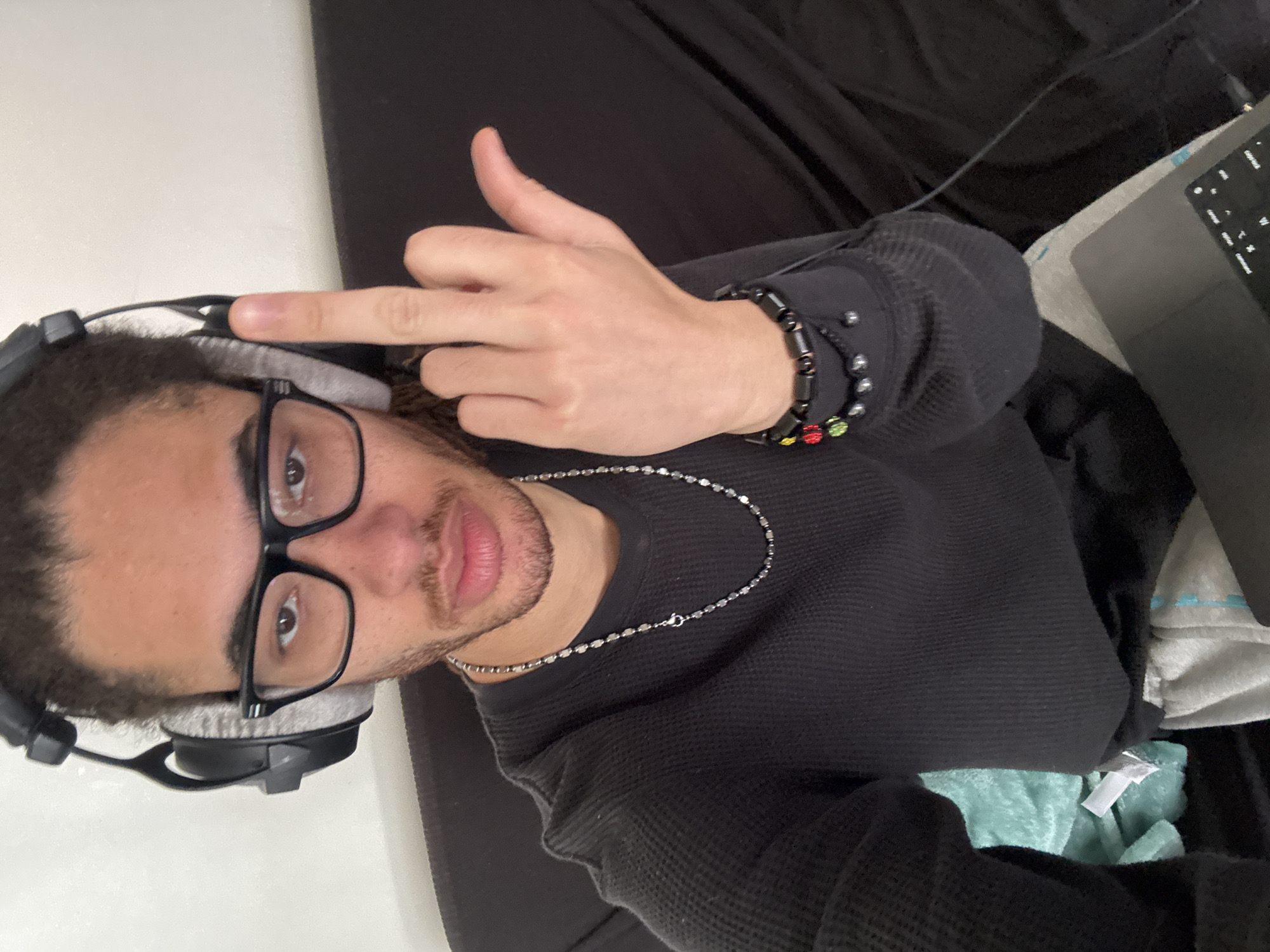 A person with dark curly hair, wearing black-rimmed glasses and headphones, is pictured from a high angle. They are wearing a black long-sleeved shirt, a silver chain necklace, and bracelets on their right wrist. Their right hand is raised, displaying the middle finger. A laptop is partially visible in the upper right corner of the frame. The background consists of dark material, likely clothing or upholstery. The image does not contain information to determine location in Paris.