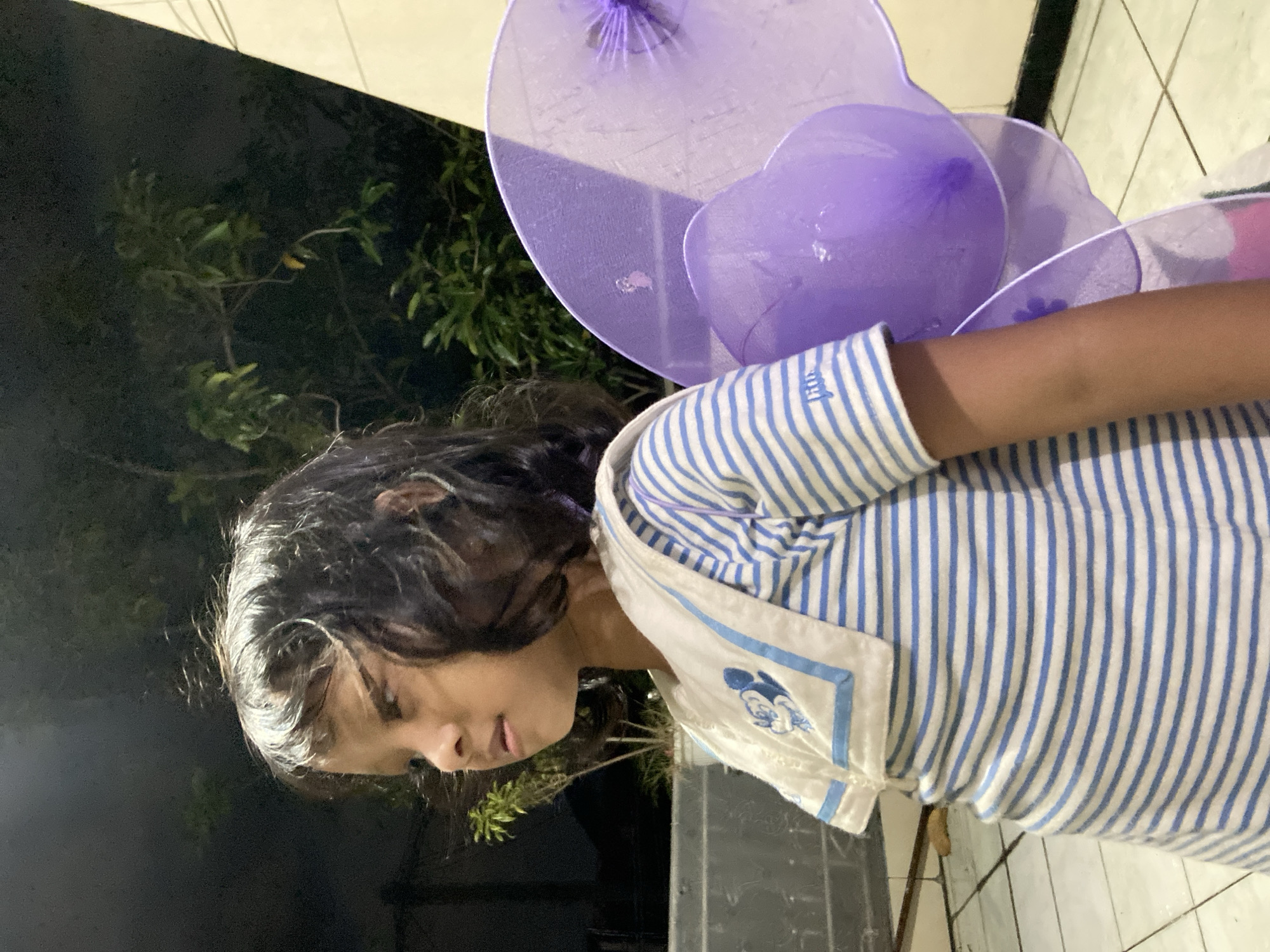 A girl is standing outdoors in Gelam, Indonesia. She has dark hair and is wearing a blue and white striped shirt. Behind her are large, purple fairy wings. The background includes a tree and a building. The lighting suggests it is either dusk or night.