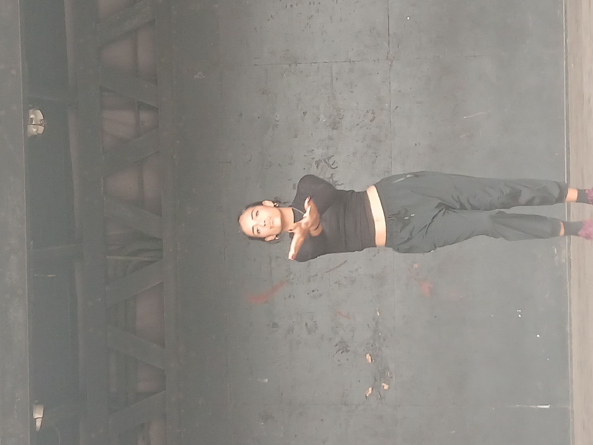 A medium shot depicts a person lying diagonally across a dull gray surface that appears to be a stage or floor. The person, facing the camera, is wearing a black long-sleeved shirt and dark pants. Their hands are crossed in front of them. The background is a muted gray, showing some markings. The left side of the image displays structural beams. The location of this image is unknown. There is no information about the city of Playa El Agua, Venezuela.