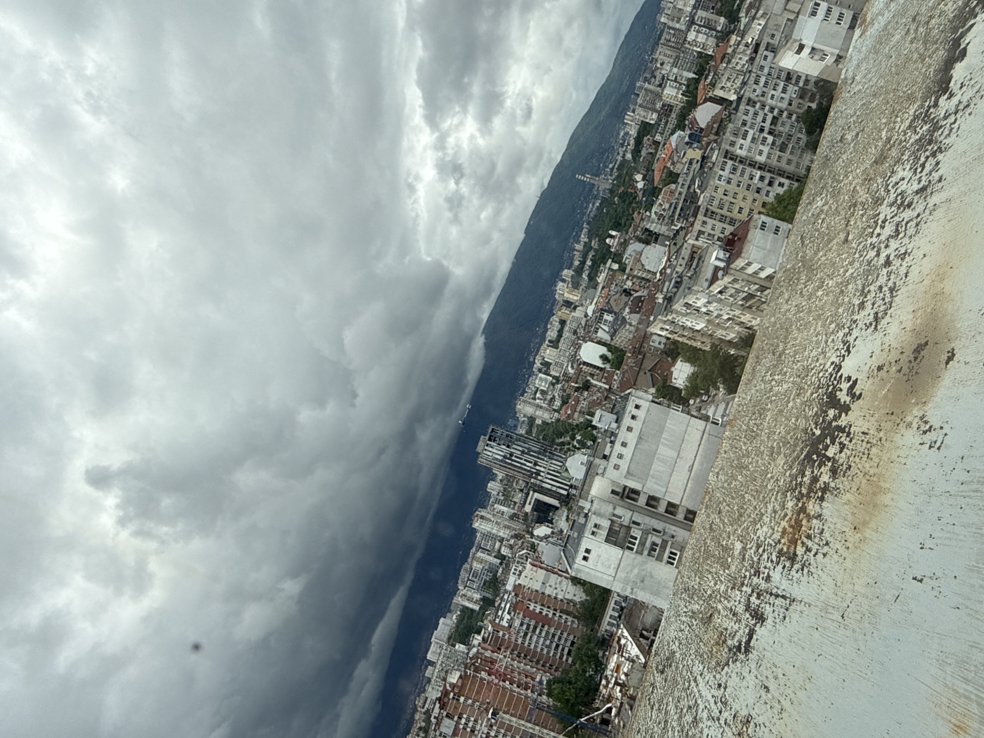 This high-angle, diagonally oriented photograph presents a sprawling urban landscape set against a dramatic, cloudy sky and a mountainous backdrop. The image appears to be taken from a significant height, possibly from a building or observation point, looking down over the city.
The sky, occupying a large portion of the upper and central frame, is filled with heavy, grey clouds, indicating overcast weather, possibly before a storm. These clouds exhibit variations in density, with darker, more ominous sections transitioning into lighter, diffused areas.
A distinct diagonal line separates the cloud-filled sky from a dark, tree-covered mountain or large hill that extends across the middle of the frame. At the base of this mountain, a dense city unfolds, characterized by numerous buildings of varied heights and architectural styles. Many structures are light-colored, featuring multiple windows, suggesting a mix of residential and commercial properties. The urban area stretches extensively, following the contours of the landscape.
In the immediate foreground, on the right side, there is a textured, light-colored surface, likely a wall, ledge, or roof edge. This surface shows considerable weathering, with dark, irregular streaks and patches that appear to be dirt, grime, or possibly rust, indicating exposure to the elements.
No people or discernible activities are visible. The time of day is daytime, under heavily overcast conditions, contributing to a somewhat somber or dramatic atmosphere. This scene is identified as Sofia, Bulgaria, with the mountain likely being part of the Vitosha massif. No visible text is present in the image.