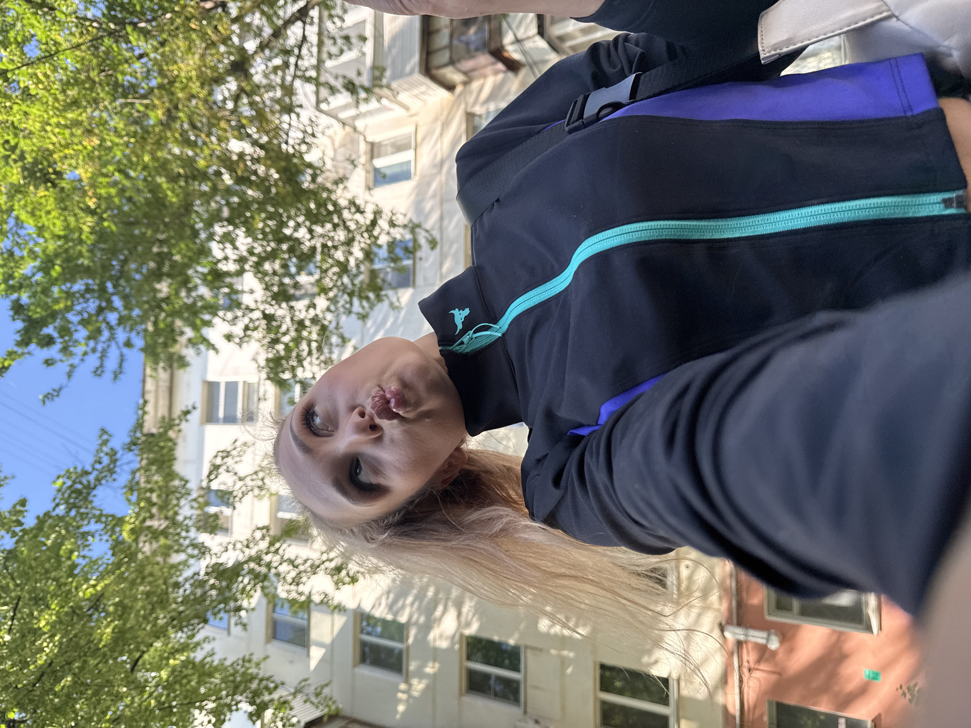 A young woman with blonde hair is looking up at the sky, puckering her lips. She is wearing a black jacket with a turquoise zipper and purple accents. A grey bag is visible on her right shoulder. Behind her, a white apartment building is visible, with trees and a blue sky overhead. The scene appears to be set in an urban environment, likely in Chaikovsky, Russia, as indicated by the architectural style of the building and the presence of trees common to the region.