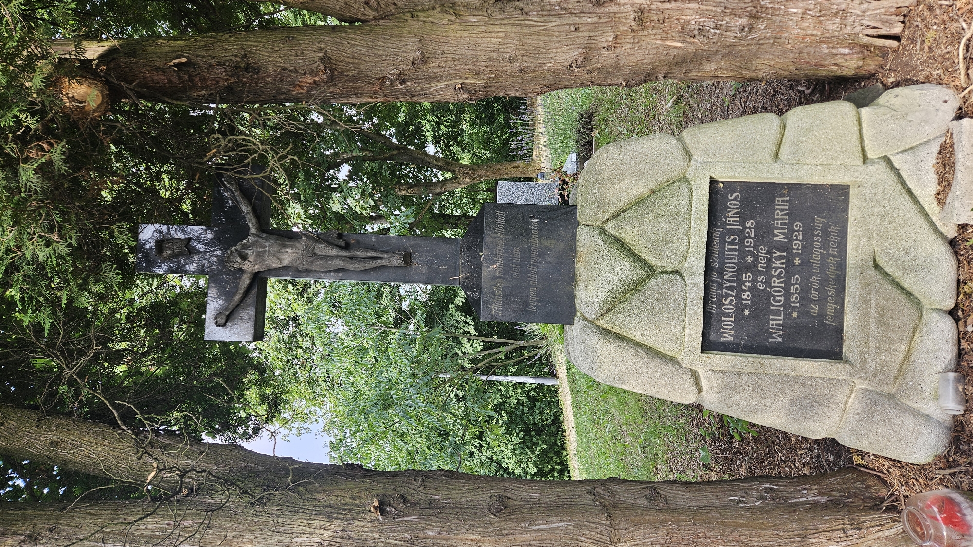 This is a medium-shot, eye-level, outdoor photograph of a crucifix monument in a cemetery. The monument is made of dark stone and depicts Jesus Christ on the cross. The base of the monument is made of rough, light-colored stones. A black plaque with white text is affixed to the base, bearing inscriptions in Hungarian. To the left and right of the monument are mature trees with thick, textured bark. The background consists of lush green foliage and trees, suggesting a natural, park-like setting. The ground is covered in grass and some scattered leaves. The overall lighting indicates a daytime scene. This monument is located in the Catholic Cemetery in Prešov, Slovakia.