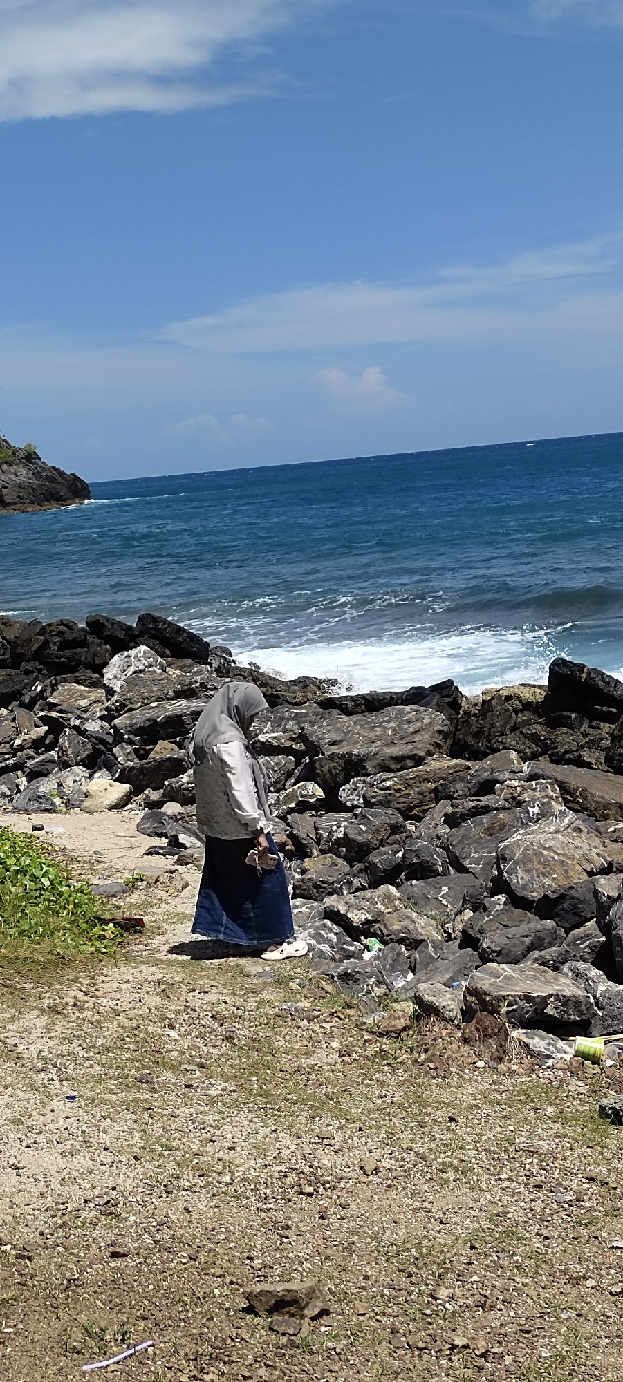 In the city of Naga Umbang, Indonesia, a lone individual wearing a grey hijab and a long blue skirt stands on a rocky shoreline. The person is holding a small object, possibly a phone, in their right hand. The foreground consists of a gravelly path with sparse green vegetation and scattered rocks. Behind the individual, a vast expanse of the ocean meets the shore, with waves crashing against the dark, rugged rocks that form the coastline. The sky above is a clear blue with a few wispy white clouds, suggesting a bright, sunny day. The overall scene depicts a serene, natural environment with a solitary figure contemplating the seascape.