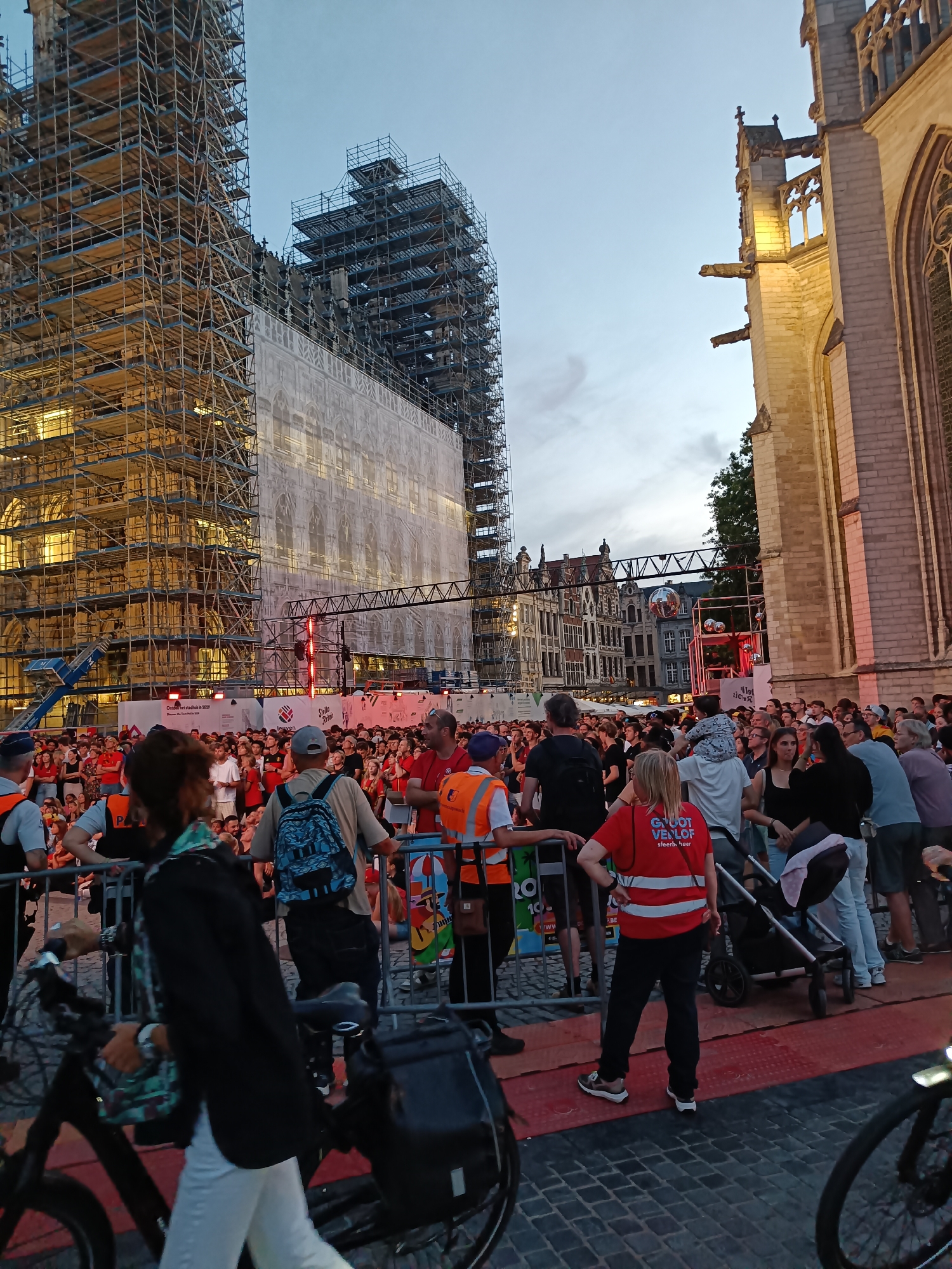 In Leuven, Belgium, a large crowd gathers outdoors in front of a grand, old building covered in scaffolding and a large white tarp. The building appears to be undergoing renovation or construction. To the right of this structure is the illuminated stone facade of another historic building, possibly a church, with gothic architectural elements. The sky is a dusky blue, indicating it is likely early evening or late twilight.
The crowd is dense, with people of various ages. Many are wearing casual clothing, and some are wearing red tops, suggesting a possible event or affiliation. In the foreground, a person on a bicycle with a basket is moving from left to right, partially obscuring the view of some individuals. Several metal barriers are set up to manage the crowd. Behind the barriers, people are standing and watching the activity. A stroller is visible amongst the crowd. In the background, beyond the scaffolding, other buildings with distinctive architecture can be seen. The overall scene suggests a public gathering or festival.