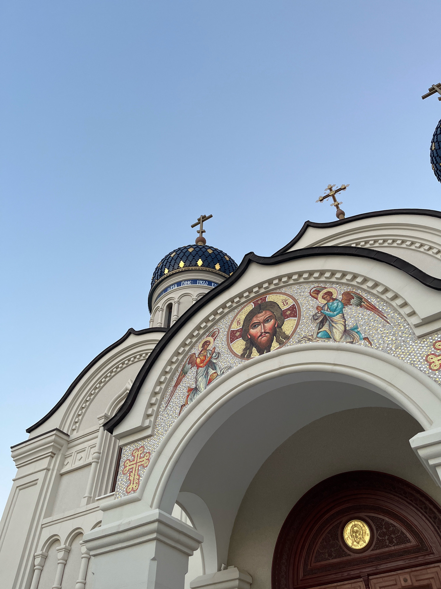 The image captures the exterior of an Orthodox Christian church with a clear blue sky as the backdrop. The building features a white facade with decorative elements and mosaics. Prominently visible are several domes topped with crosses. One of the domes is dark blue with gold accents and inscribed with Cyrillic lettering. The mosaics depict religious figures, including a representation of Jesus Christ and angels, along with various crosses. The architecture includes arched entrances and decorative cornices. The overall scene is one of religious architecture bathed in natural light. This church is located in the municipality of Germasogeia, Cyprus.