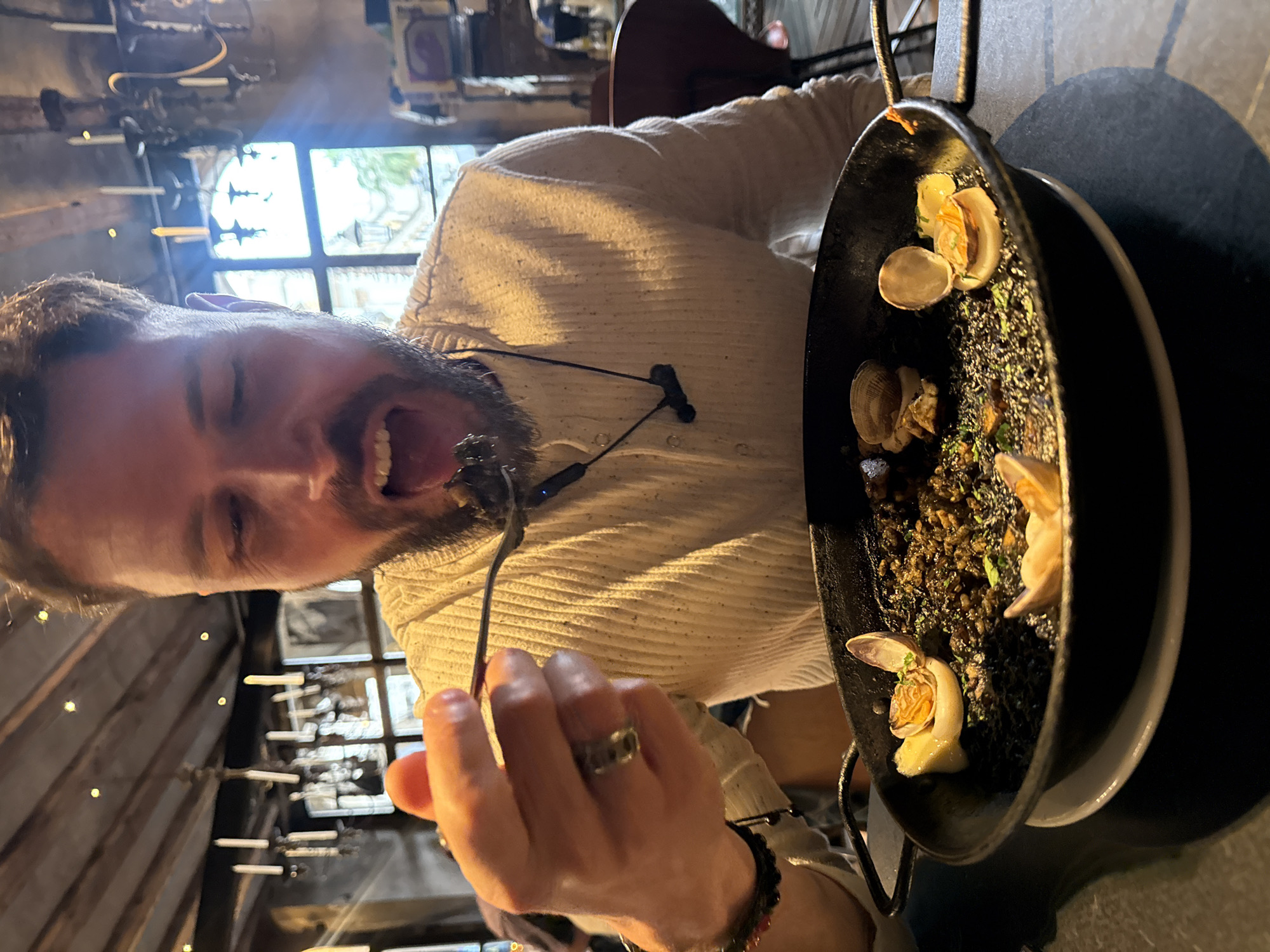 A man is eating a meal, possibly black paella, from a black pan. The pan is set on a black table. The man is holding a forkful of the meal near his mouth. He is wearing a light-colored ribbed shirt and has a black wireless headset around his neck. He is also wearing a ring and a beaded bracelet.
The background shows a window and parts of what looks like a rustic interior with candle-like fixtures. This image was taken at a restaurant called 7 Portes in Barcelona, Spain.