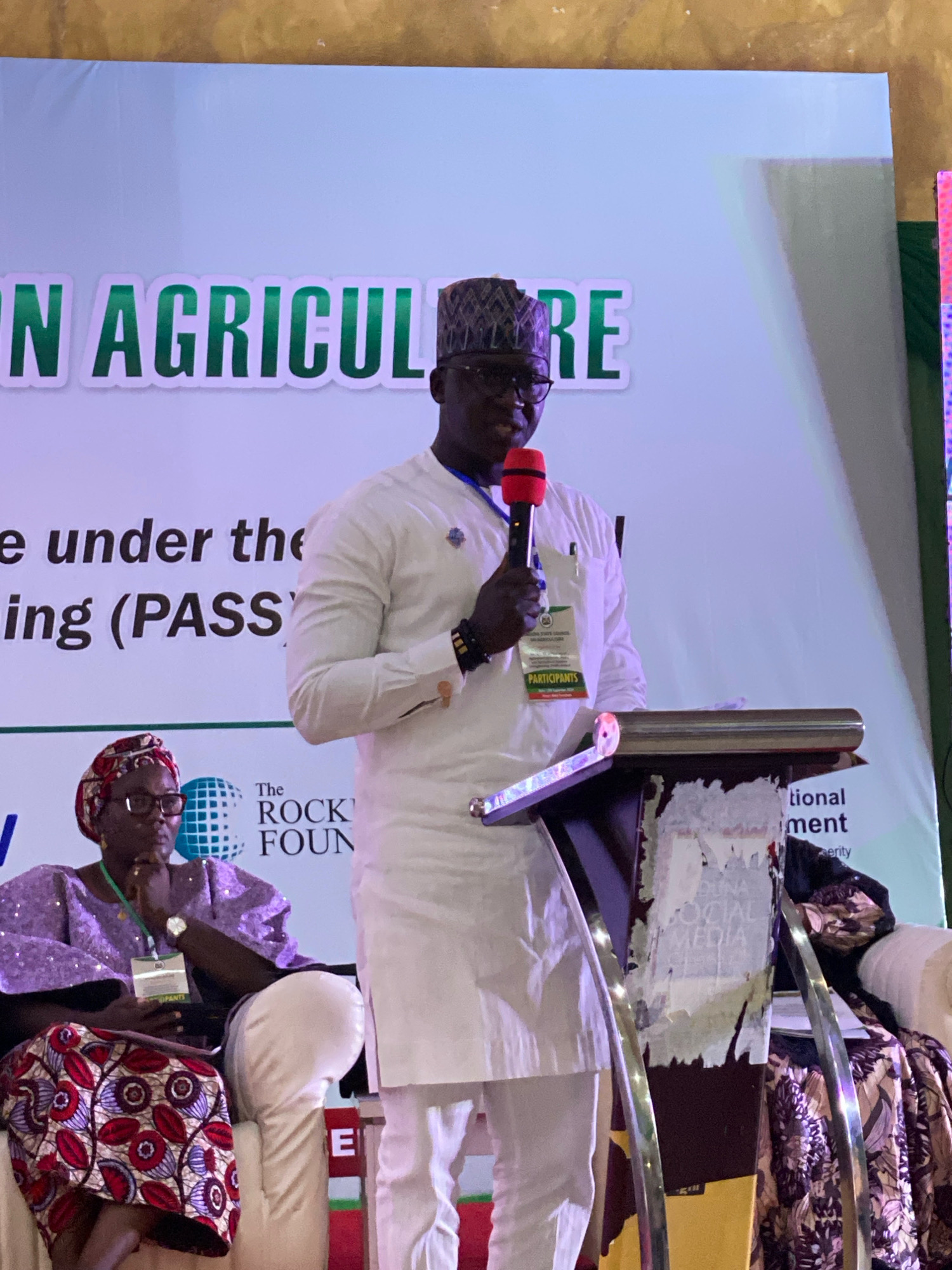 The image depicts an indoor event with a light blue backdrop featuring green lettering that reads "N AGRICULTURE". A Black man wearing a white traditional outfit and a patterned cap is speaking into a red microphone, holding it with his right hand. He has glasses on and a badge clipped to his shirt. To his left, a Black woman is seated, wearing a purple sequined top and a headscarf with a red and white patterned print. She is also wearing glasses and a watch. Behind the woman, the words "The ROCK FOUN" are visible on a white banner. A podium with metal accents is positioned in front of the speaker. The overall environment suggests a conference, seminar, or similar public gathering. The text "SOCIAL MEDIA" is visible on a torn poster attached to the podium.