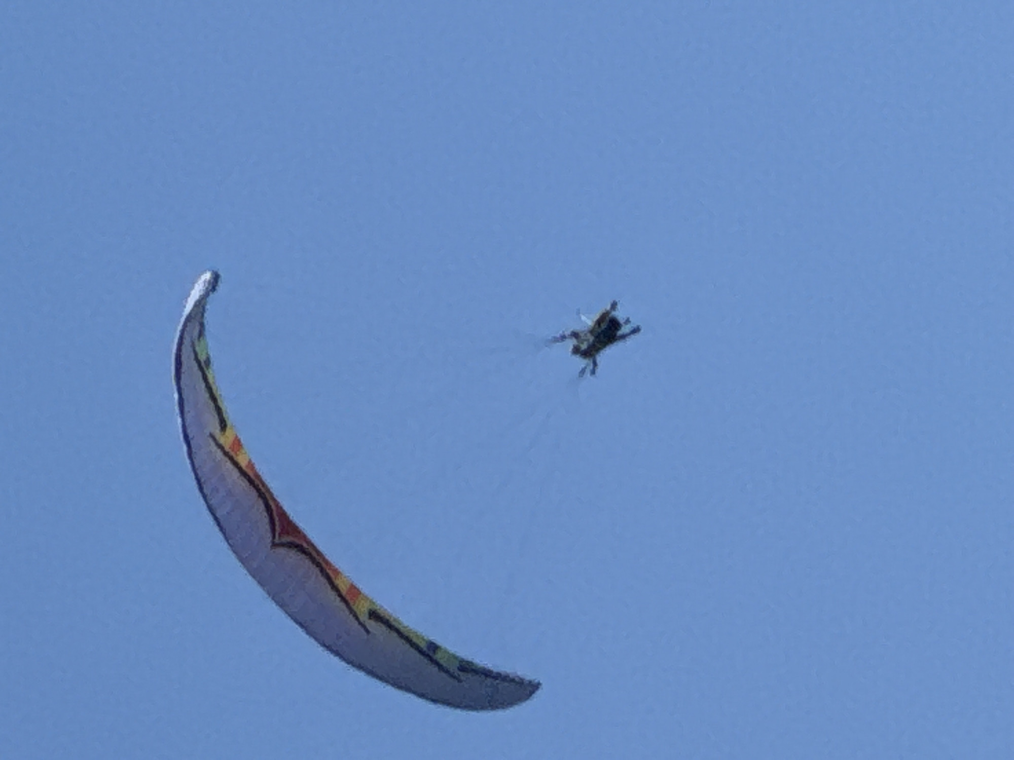 An extreme telephoto, low-resolution, overexposed, full-shot, outdoor, daytime, vertical image depicts a paraglider in mid-air against a clear blue sky. The paraglider, a colorful canopy with stripes of red, yellow, and blue, is positioned in the upper left quadrant of the frame. Suspended beneath the canopy, a small, dark figure, presumably the paraglider, is visible. The paraglider appears to be descending or maneuvering. The background is a uniform, bright blue sky with no discernible landmarks or features. The image quality is poor, with significant pixelation and noise, obscuring fine details.