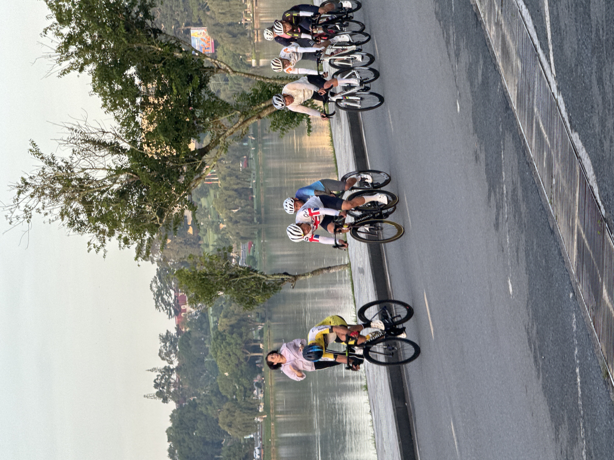 In Phường Xuân Hương - Đà Lạt, Vietnam, a group of cyclists, dressed in athletic gear and wearing helmets, are racing on a paved road. The cyclists are positioned in the mid-ground of the image, with several of them riding in a pack. To the left of the cyclists, a lone woman in a pink jacket is running alongside the road, facing the cyclists. The background features a large body of water, likely Xuan Huong Lake, with trees and distant buildings visible along its shore. The sky is overcast, suggesting an early morning or late afternoon setting. The road is marked with white lines, and a metal grate runs along the edge of the pavement in the foreground.