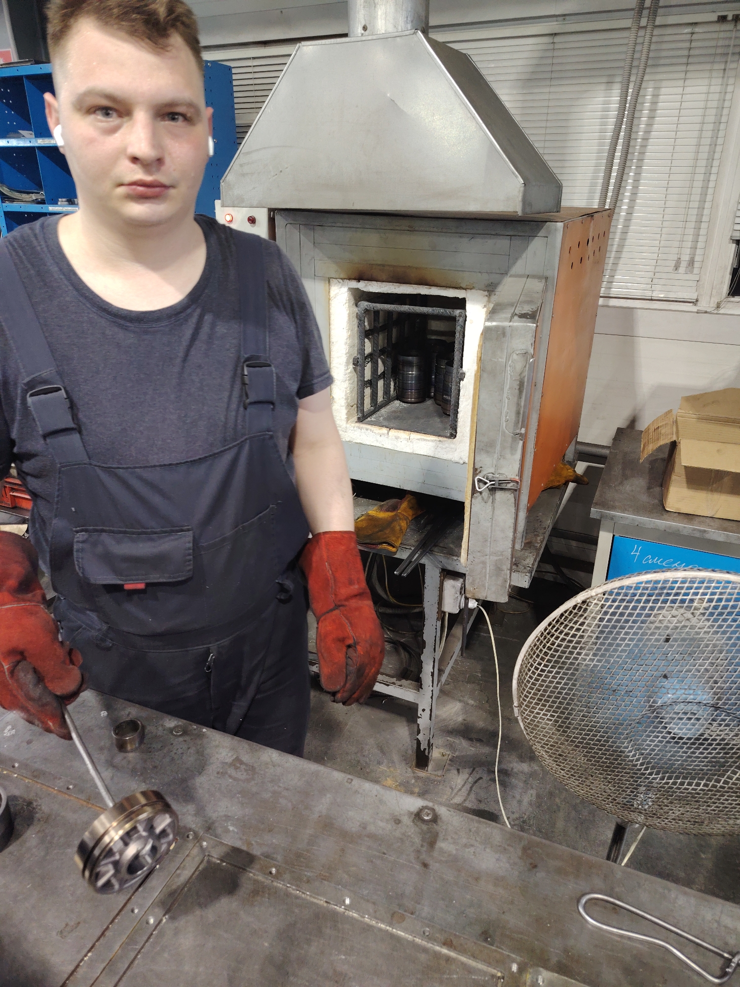 A man in dark-blue overalls, wearing wireless earbuds and red heat-resistant gloves, stands in what appears to be a workshop, operating an industrial furnace. The furnace is open, revealing metal objects inside. The man holds a long metal rod with a metal object at the end. A large fan sits on the floor to his right. A blue shelf unit is partially visible in the upper left corner. The image has a utilitarian, industrial feel. There is no indication the location is Noviy, Russia.