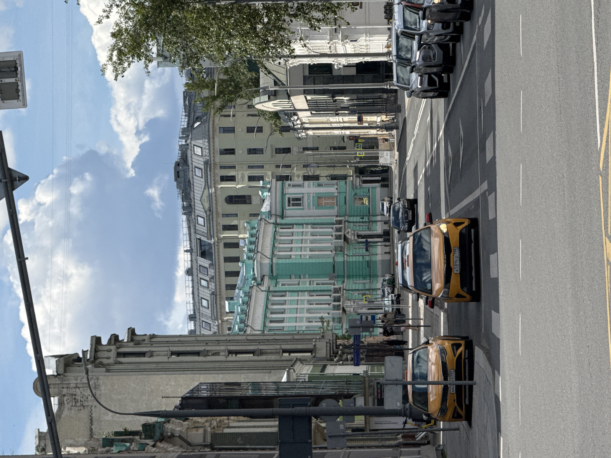 This is a street-level view of a city street in Moscow, Russia, on a partly cloudy day. The perspective is from the middle of the street, looking forward. Two yellow taxi cabs are prominent in the foreground, parked on the right side of the road. The one on the left has license plate M444TT 792 and the one on the right has license plate A589EE 799. They are both facing away from the viewer.
The street is lined with multi-story buildings. On the left, there is a light-colored, ornate building with arched windows and decorative architectural elements. Further down the street, on the left, is a striking turquoise and white building with a very structured facade. On the right side of the street, the buildings are lighter in color, with more traditional window designs and some trees growing alongside the sidewalk.
Further down the street, traffic can be seen. Several cars are parked or moving. Pedestrians are visible on the sidewalk, walking past the buildings. Traffic lights and street signs are present along the route. The sky is blue with dramatic white clouds. The overall impression is of an urban environment with a mix of older and more modern architecture.
