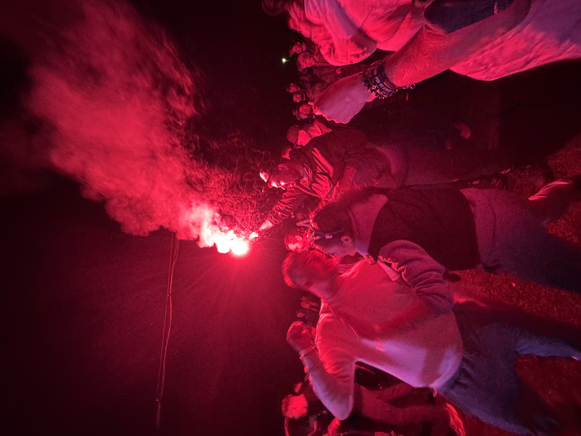 The image depicts an outdoor nighttime gathering of multiple individuals in Большая Кивара, Russia, illuminated predominantly by the intense red light and smoke emitted from a handheld flare. A male individual positioned towards the right of the frame holds the actively burning red flare aloft in his right hand, generating a thick plume of red smoke that dissipates into the dark sky. This illumination casts a strong red hue over the entire scene and the attending crowd.
In the mid-ground, to the left of the flare-holder, another male individual wearing a light-colored sweatshirt is visible, with his left arm raised or bent, his body oriented towards the activity. Further left and partially obscured, additional figures are present. In the lower-left foreground, a person in a light top and dark pants is in motion. In the lower-right foreground, a raised arm adorned with multiple dark, possibly beaded bracelets, is visible. The background reveals a dense crowd of people, mostly silhouetted or dimly lit by the red light, indicating a significant number of participants. Overhead in the upper-left, faint horizontal lines, possibly wires or cables, are discernible against the dark sky. The ground beneath the individuals appears to be an uneven outdoor surface.