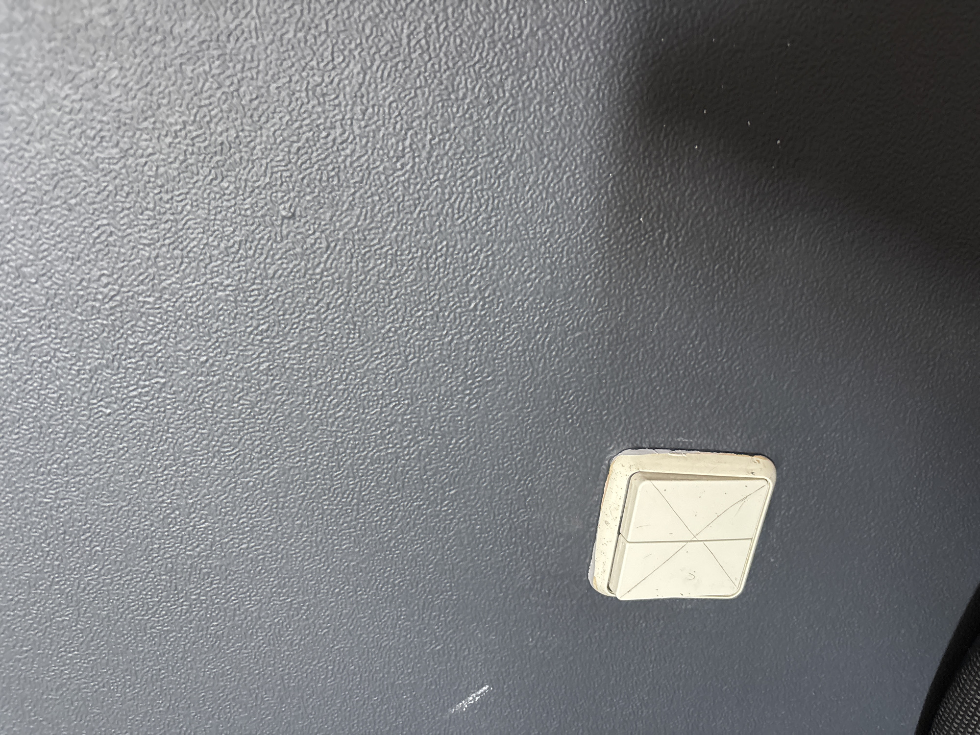 An indoor shot shows a textured gray wall with a single cream-colored light switch. The switch is square with a prominent beveled edge, and its surface is divided into four triangular sections by lines etched into the plastic. The switch appears to be older, with some scuff marks and minor discoloration around the edges where it meets the wall. A faint white scratch is visible on the wall to the left of the switch. The lower left corner of the image is partially obscured by a dark, textured object, possibly a bag or piece of furniture. The lighting suggests an indoor environment with ambient light. The location of this scene within Zaporizhzhia, Ukraine is not determinable from the image itself.