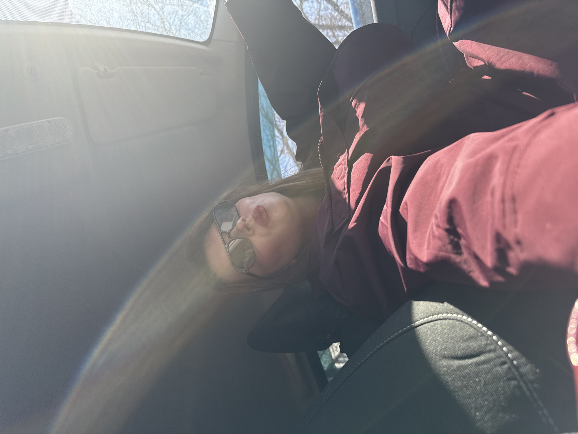 The image captures a person reclining inside a car, viewed from a low, tilted angle. The primary subject is a person, likely female, with long brown hair, wearing dark sunglasses and a dark maroon or deep red hooded jacket. They appear to be looking upwards or slightly away from the camera, with their face partially obscured by the sunglasses and strong sunlight.
The scene is set inside a car, with elements of the vehicle's interior visible, including the light-colored overhead ceiling, a sun visor, and part of a dark grey car seat with white stitching in the foreground. Strong sunlight streams into the car from the upper left, creating significant lens flare and a visible rainbow light refraction across the frame. This intense backlighting casts much of the car's interior and parts of the person in shadow, while highlighting textures on the jacket and seat.
Through the car windows, the background shows bare tree branches against a bright, hazy sky, suggesting a winter or early spring setting in Izhevsk, Russia. The sunlight indicates it is daytime. No specific activity beyond resting or observing the surroundings is discernible. There is no visible text in the image. The overall mood is relaxed, despite the striking lighting effects.