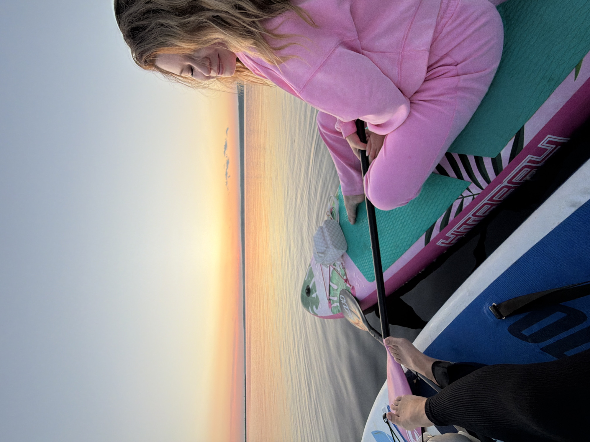 The image shows two people on paddleboards on a calm body of water, likely at sunset. The person in the foreground is a young woman wearing a pink velour tracksuit, sitting cross-legged on a paddleboard. She has her eyes closed and appears relaxed, with her blond hair framing her face. Her paddle is resting on the board in front of her. Beside her, on another paddleboard, are the feet of another person wearing black leggings. The paddleboards are colorful, one with pink and green accents and the other predominantly blue and white. The water is still, reflecting the soft hues of the sunset, which paints the sky in shades of orange, pink, and light blue. The horizon is visible in the distance, with a faint outline of land. The location is identified as the city of Chaikovsky, Russia.