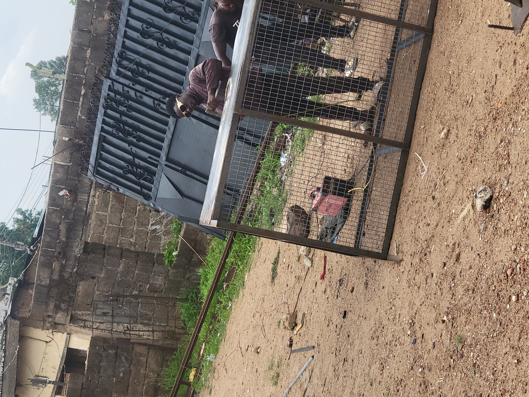 In Jos, Nigeria, the image shows two individuals working on a metal structure. One man, wearing a maroon shirt and distinctive sunglasses, is actively engaged in working with a metal gate or frame. A second person is partially visible behind the structure. The metal structure appears to be a cage or frame made of metal bars and wire mesh. The setting is a dusty, sandy area with scattered debris. In the background, a weathered brick building is visible, along with a section of decorative metal gates.