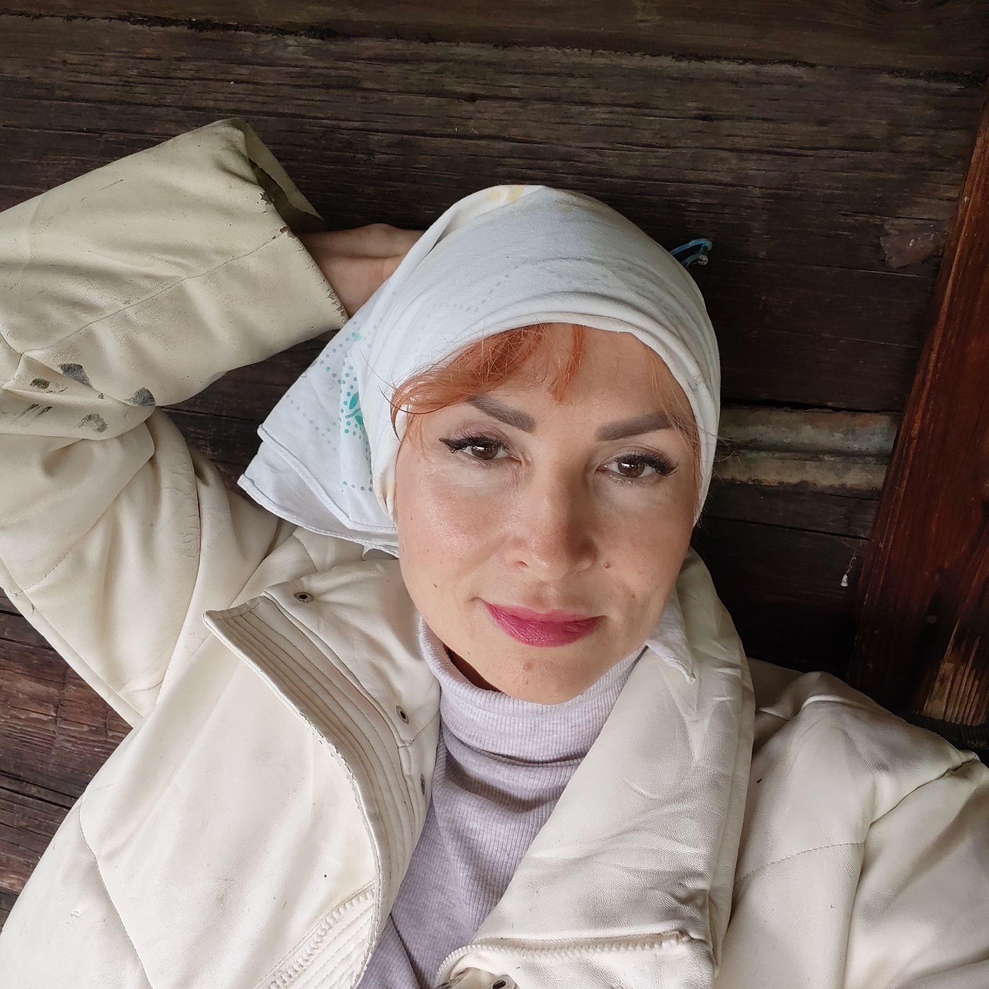 This image captures an adult female posing for a photograph in the city of Chaikovsky, Russia, against a backdrop of dark, weathered wooden planks. The woman, estimated to be in her 40s or 50s, is positioned in a reclining manner, looking directly at the camera with a slight smile. She wears a white headscarf with a subtle light blue dotted and floral pattern, revealing some reddish-brown hair beneath. Her makeup includes dark winged eyeliner, possibly false eyelashes, and bright pink lipstick. Her skin shows a few visible moles or freckles. She is dressed in a light purple ribbed turtleneck sweater and an off-white or cream-colored padded jacket, which exhibits wear such as dark smudges and scuffs, particularly on the sleeves and collar area. Her right arm is bent and positioned behind her head, providing support. The wooden background consists of aged, textured planks with visible grain and minor gaps, suggesting an interior or sheltered rustic environment.