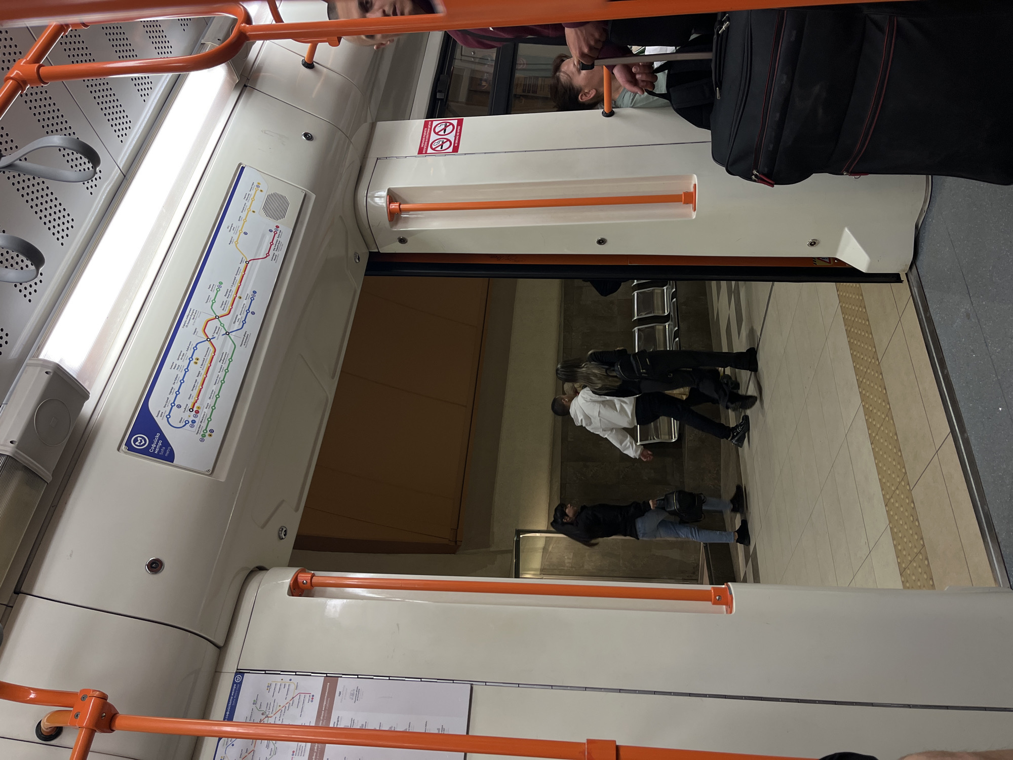 The image captures the interior of a metro train, looking out onto a station platform, rotated 90 degrees clockwise. Inside the train, prominent orange handrails and grey hanging straps are visible against white and grey perforated panels. Overhead, long fluorescent lights illuminate the carriage. A metro map, featuring a stylized "M" logo and several colored lines (red, green, blue, yellow, orange), is displayed on the wall. Partially legible text on the map includes "Oborishte Kertsvo". A "No Smoking" sign, depicted as a red circle with a crossed-out cigarette, is affixed near the door. Reflections of people inside the train, including a person's face at the top and a woman's head near the door, are visible in the glass. A large black suitcase is also inside the train, near the exit.
On the station platform, a tiled floor with distinct tactile paving strips for the visually impaired is visible. Several people are standing or walking on the platform: one individual wears a white jacket, another a black top and light blue jeans, and the lower body of a third person is also seen. The platform appears moderately lit, characteristic of an underground station. The scene depicts a typical metro environment in Sofia, Bulgaria.