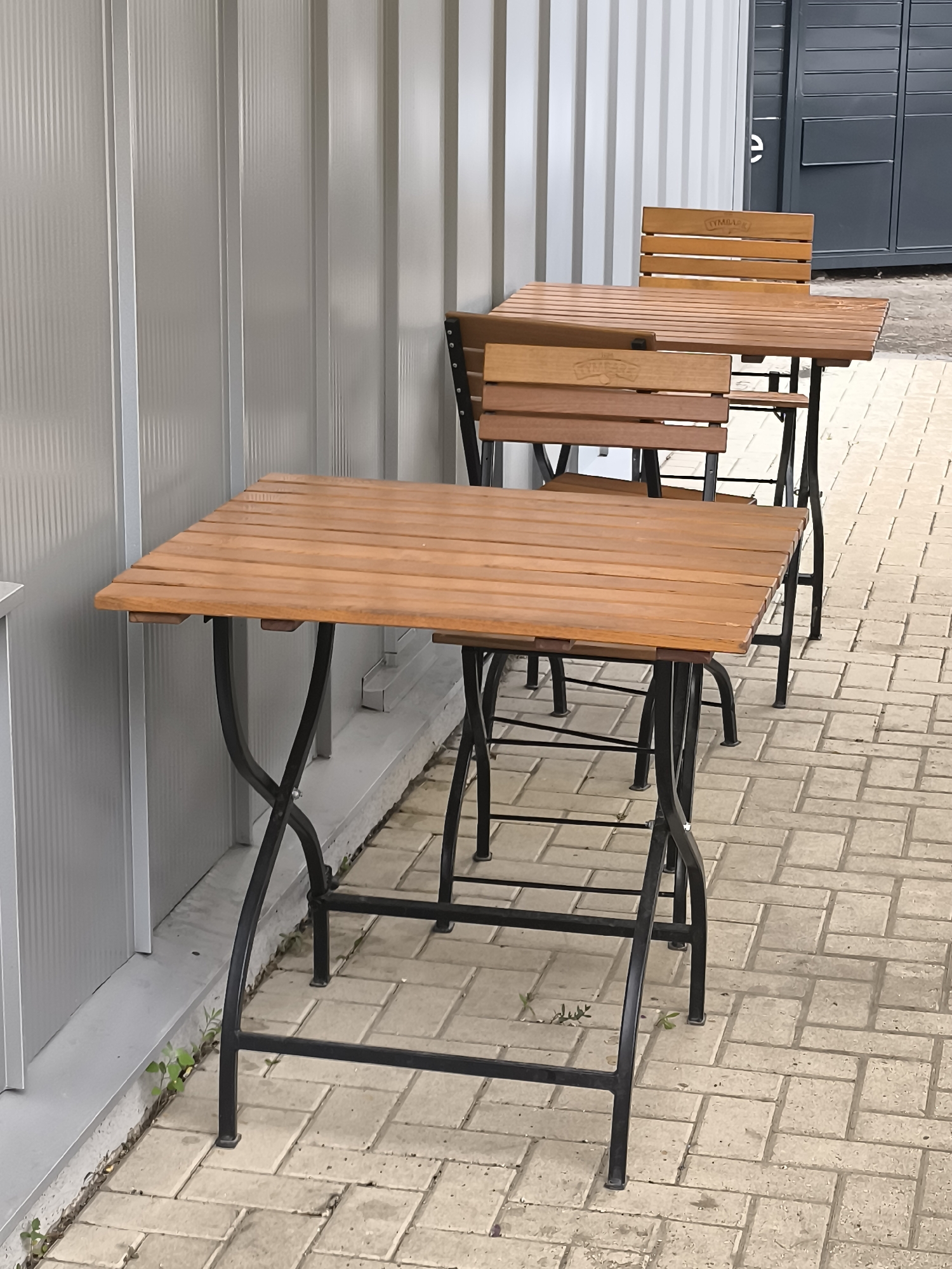The image depicts an outdoor seating area, likely an extension of a business, featuring two wooden tables with black metal frames and matching wooden chairs. The tables are positioned on a paved walkway, composed of rectangular light-colored interlocking bricks. A corrugated metal wall forms the backdrop to the left of the scene. To the right, a dark grey modular structure with visible doors is present. The setting appears to be a public space within the city of Bălți, Moldova. There are no people visible in the image. The tables and chairs are neatly arranged, suggesting a functional outdoor dining or seating arrangement.