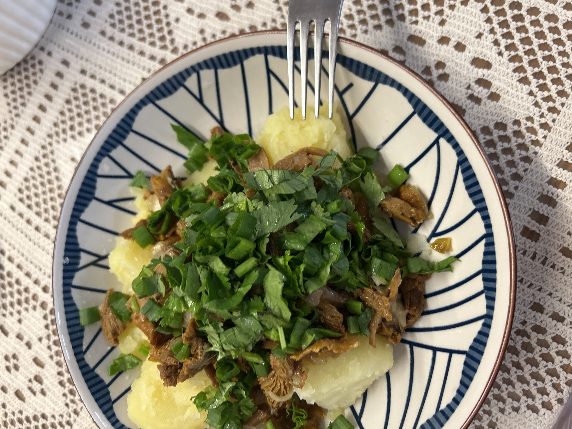 A top-down shot captures a blue and white patterned plate filled with what appears to be a homemade dish. The main components of the dish are boiled potatoes, shredded meat or mushrooms, cilantro, and scallions. A silver fork pierces one of the potatoes. The plate rests on a white lace tablecloth. The location cannot be determined from the image.