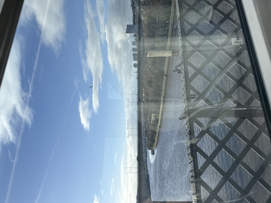 A high-angle view from a window showcases a landscape featuring a river winding between grassy banks, a city skyline in the distance, and a bright blue sky with scattered clouds. The architectural framework of a bridge dominates the right side of the image, while a bird soars freely in the sky. The scene is bathed in natural light, creating a tranquil and expansive vista.