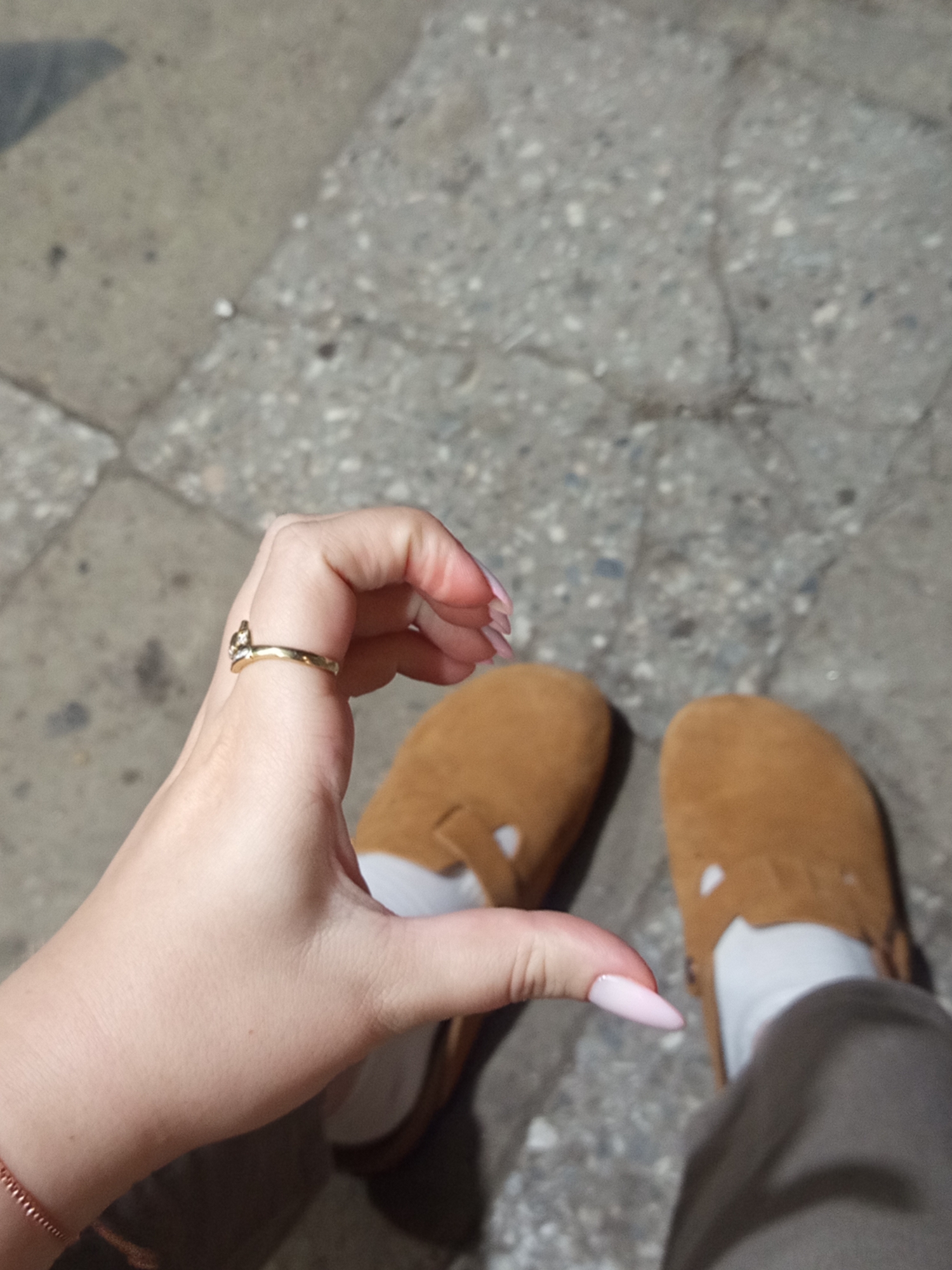A close-up, waist-up shot shows a person's hand forming a heart shape with their thumb and index finger, with the palm facing the camera. The hand has long, light pink acrylic nails. A thin, gold ring with a small stone is visible on the index finger. The person is wearing a simple brown bracelet on their wrist. In the background, the person's feet are visible, wearing tan, slip-on shoes with white lining and white socks. The shoes are resting on a cracked, grey concrete surface, typical of a sidewalk or paved area. The overall lighting suggests an outdoor setting, possibly in the city of Khashuri, Georgia.