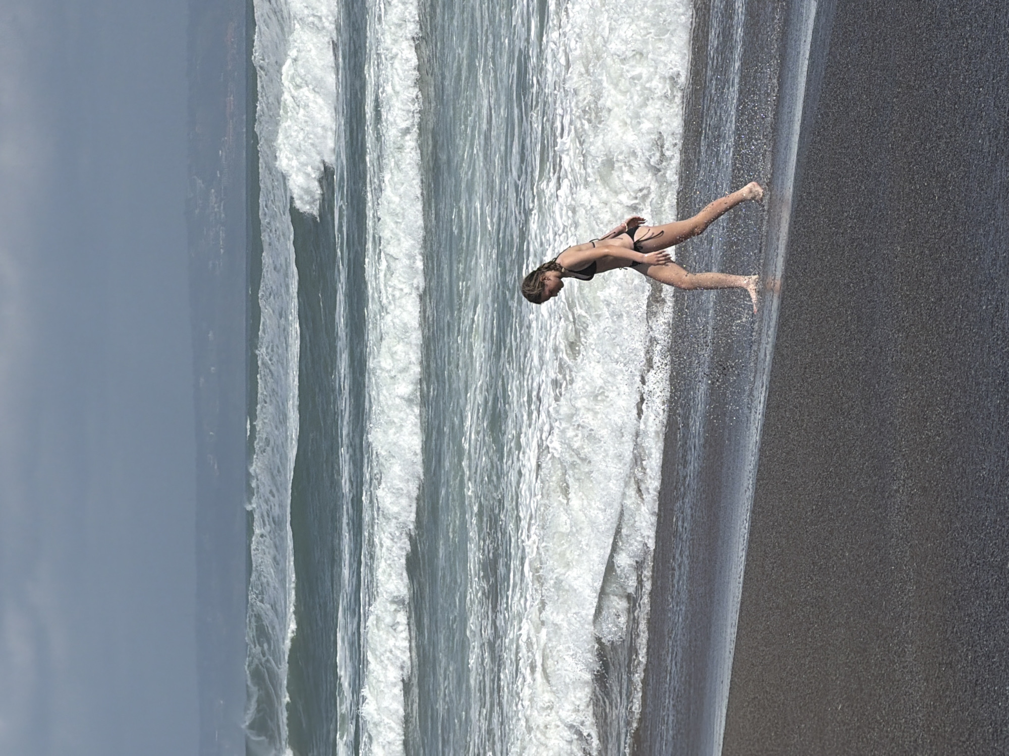 A young woman in a black bikini walks along a dark sand beach in North Kuta, Indonesia. She is facing to the left, with her right leg forward and her left leg trailing. The ocean is to her right, with large waves crashing onto the shore. The sky is overcast and the water is a murky green color. In the background, there are hazy hills that suggest a distant coastline.