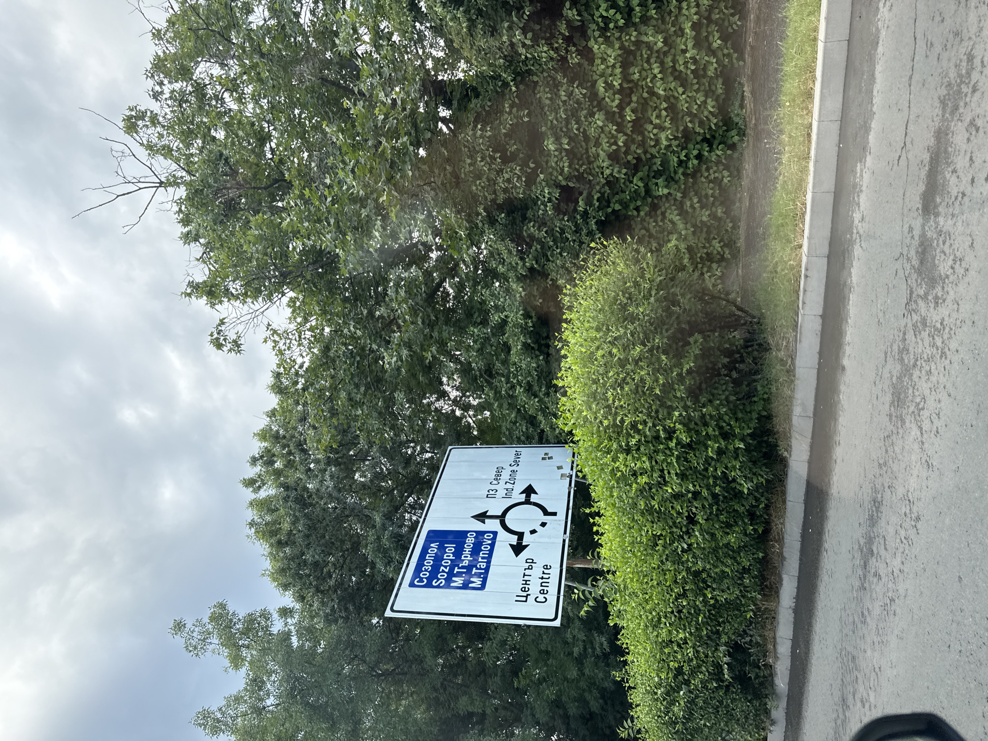 The media file displays a roadside scene in Burgas, Bulgaria, under an overcast sky. The primary subject is a blue and white road sign providing directional information. The sign indicates routes to "Sozopol", "M.Tarnovo", and "Centre" in both Bulgarian and English. Additionally, it shows directions to "ПЗ Север" (which translates to "Industrial Zone North") and "Ind. Zone Sever", suggesting an industrial area. A circular diagram with arrows illustrates a roundabout ahead.
The setting is a paved road bordered by a concrete curb and a strip of grass. Lush green foliage, consisting of trees and meticulously trimmed bushes, forms a dense backdrop on the left side of the image. The lighting suggests it is daytime, though the cloudy weather casts a diffused light over the scene, creating a slightly muted atmosphere. There are no people visible in the image. The overall impression is one of a typical suburban or semi-industrial roadside in Bulgaria, with clear directional signage for navigating the area.