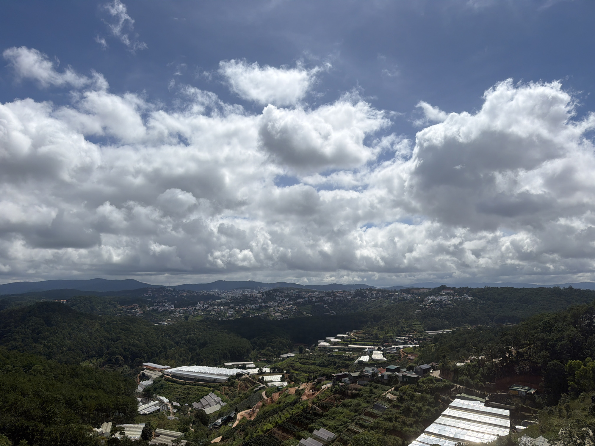 This is a high-angle, wide-shot photograph taken in the city of Xuan Truong - Da Lat, Vietnam. The scene captures a vast expanse of rolling green hills covered in dense vegetation, dotted with numerous greenhouses and some residential buildings. In the foreground, several large, white-tented structures, presumably greenhouses, are prominent on the hillside. The midground reveals more greenhouses, interspersed with smaller buildings and terraced plots of land. Further in the distance, a city sprawls across a valley, with buildings climbing up the slopes of hills. The background is dominated by a dramatic sky filled with a mixture of dark, heavy clouds and bright, puffy white clouds, with patches of blue sky visible. The overall impression is one of a rural or semi-rural area dedicated to agriculture and residential development, set against a majestic natural landscape.
