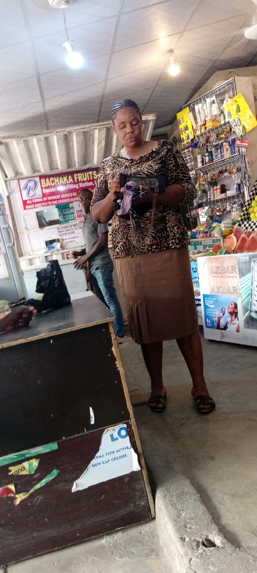 This image depicts a scene inside a shop, likely a convenience store or market stall, in Aviation Quarters, Nigeria. The setting appears to be indoors with a tiled ceiling and fluorescent lighting, suggesting it's daytime.
The primary subject is a woman with braided hair, wearing a patterned long-sleeved shirt and a brown skirt, standing and holding a small bag and a mobile phone. She is looking down at her hands. To her left, a younger man in a grey t-shirt and jeans is visible, looking towards the camera with a neutral expression. In the foreground, a dark wooden counter is visible, with a black plastic bag on it. Behind the counter, shelves are stocked with various goods, including toiletries, keys, and some fruit. A sign for "BACHAKA FRUITS" is prominent on the wall, indicating the type of business. Other visible text on signs includes "AKBAR," "LO," and "NEW CAP COLORS," suggesting a variety of products are for sale. The overall atmosphere seems to be one of everyday commerce.