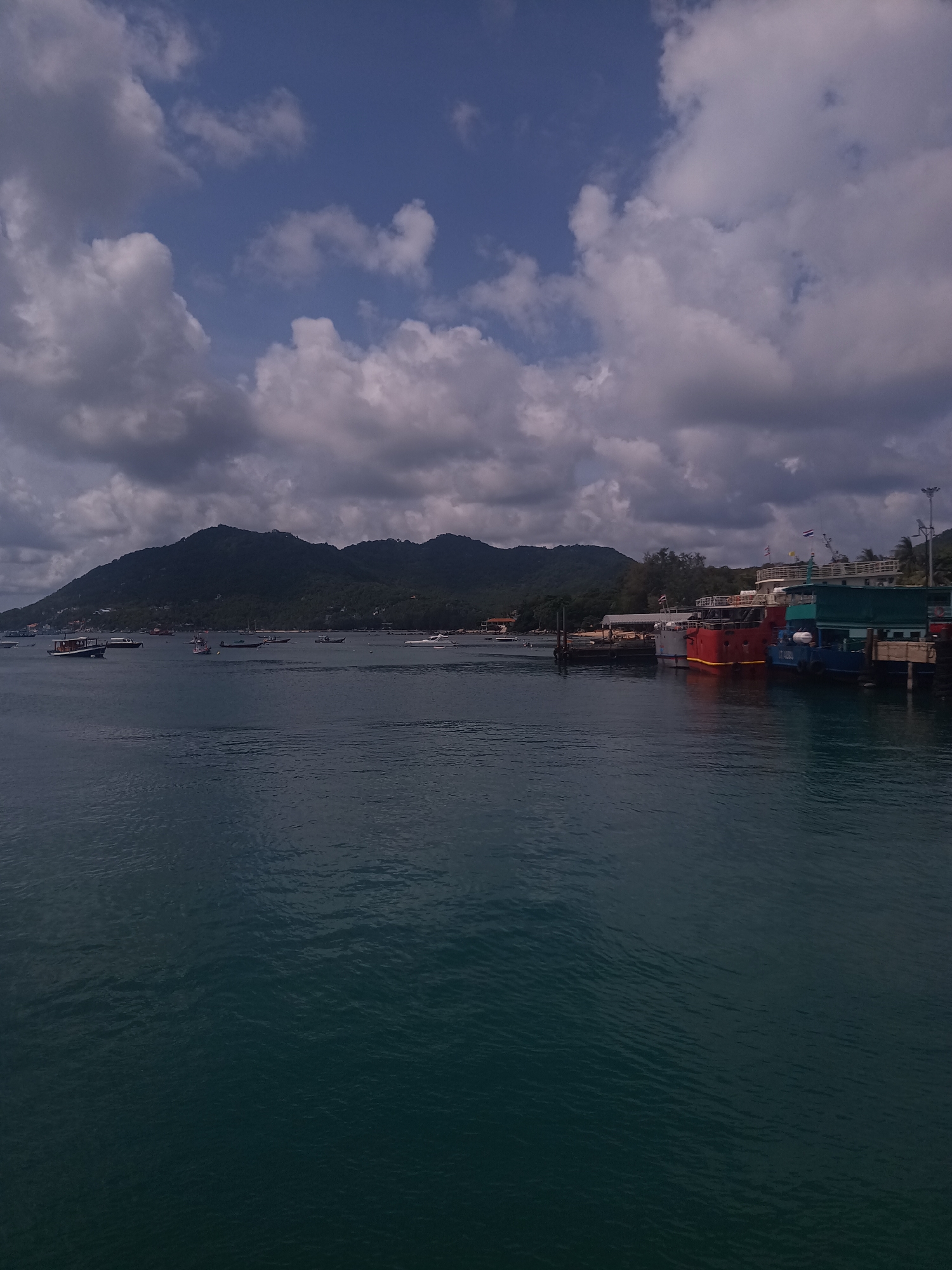 The image depicts a coastal scene in Tambon Ko Tao, Thailand. The foreground is dominated by dark turquoise ocean water. In the midground, a mountainous, forested island stretches across the horizon, dotted with buildings and structures. Multiple boats are scattered on the water's surface in the distance. To the right, a dock is visible, lined with colorful boats, predominantly red and blue. Above, the sky is partially cloudy with both dark, ominous clouds and patches of bright blue.