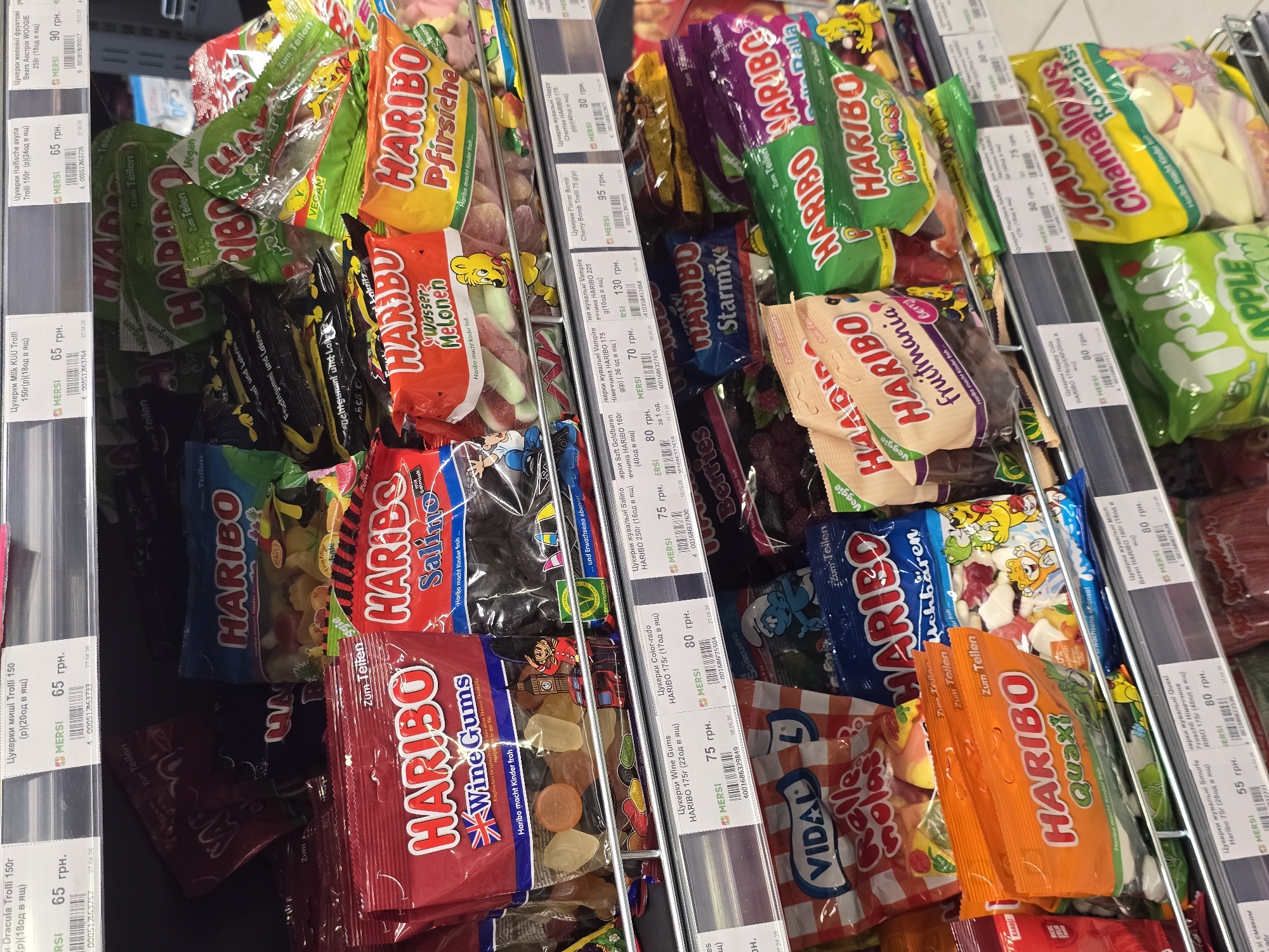 The image shows a retail shelf in a store, likely a supermarket or convenience store, stocked with various bags of Haribo gummy candies. The candies are arranged in rows on metal shelves, with price tags visible for some items. The lighting is bright and consistent with indoor store lighting. The products are predominantly bags of Haribo gummies, with labels in Ukrainian and German. Some of the visible product names include "Haribo Wine Gums," "Haribo Salino," "Haribo Wasser Melonen," "Haribo Pfirsiche," "Haribo Color-rado," "Haribo Starmix," "Haribo Quaxi," "Haribo Fruitmania," and "Haribo Chamallows Rombls." The price tags indicate prices in Ukrainian Hryvnia (грн.). The environment suggests a commercial setting focused on confectionery sales. There are no people visible in the image. The location mentioned, Petropavlivska Borshchahivka, Ukraine, is a town, suggesting this is a retail outlet within that area.