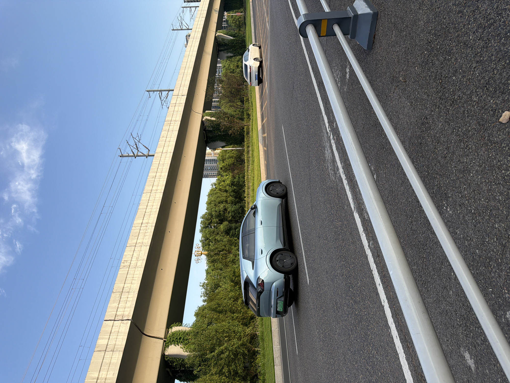 A light blue electric car is captured in motion on a paved road in Jiangjin, China, during daylight. The car, a hatchback, is positioned in the left lane and is driving towards the viewer's right. It has tinted windows and distinctive geometric wheel designs. In the right lane, a white sedan is also moving in the same direction, further away from the camera.
Overhead, a multi-lane elevated highway or viaduct stretches across the frame, supported by concrete pillars partially covered in green ivy. Power lines are strung along the top of the viaduct. The environment includes lush green trees lining the road and dense residential buildings in the background. A silver metal guardrail with yellow accents is visible in the foreground, parallel to the road. The sky is a clear blue with a few wispy clouds. The lighting suggests it is late afternoon or early morning, with soft sunlight illuminating the scene.