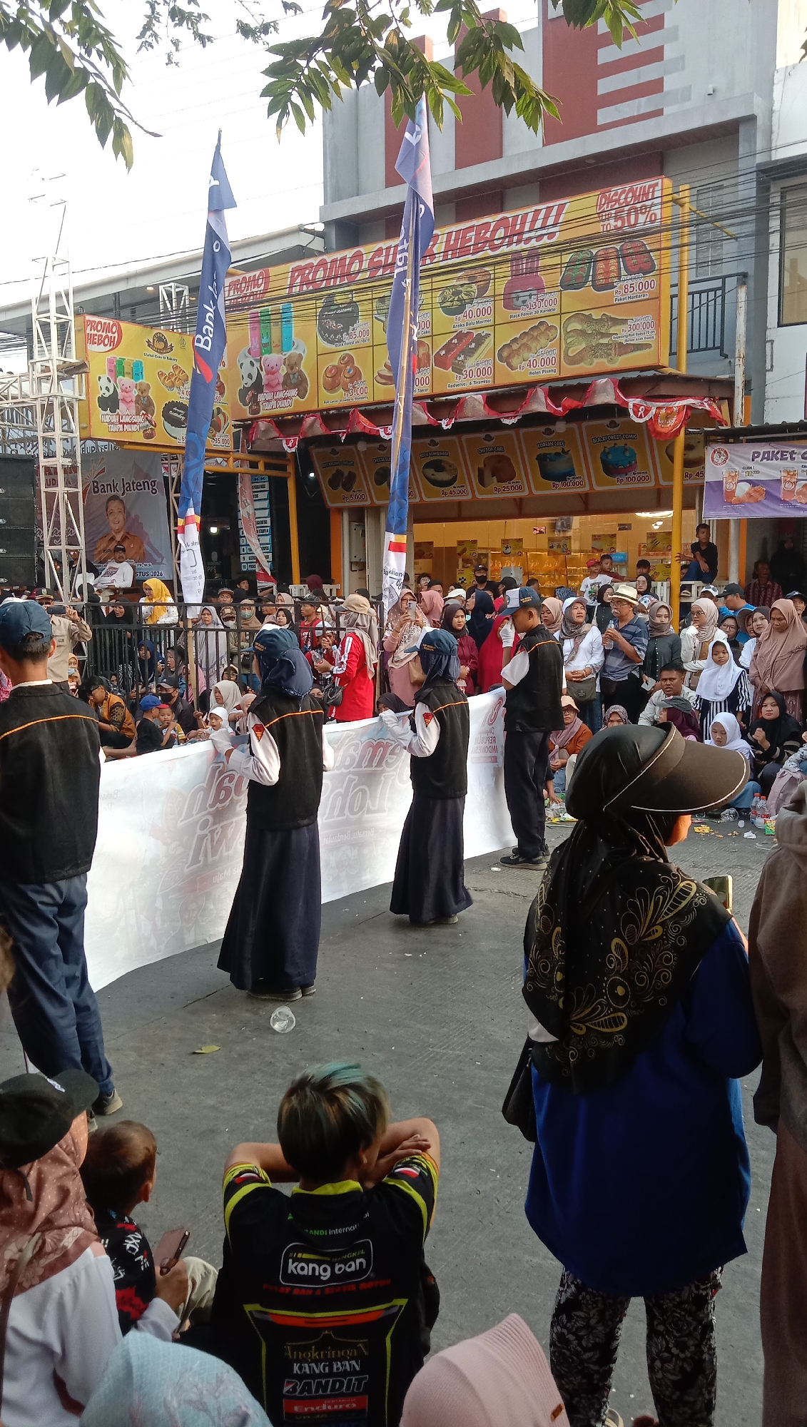 The image depicts an outdoor event in Jatisawit, Indonesia, possibly a festival or promotional gathering. In the foreground, three young individuals in dark blue abayas and vests, likely performers, stand behind a large white banner. A young person with dyed hair sits in the lower right, facing away from the camera, observing the event. Behind the performers, a crowd of people of various ages are gathered, some standing, some seated, and some watching the proceedings.
The background features a building with a red and white facade, adorned with banners advertising promotions for baked goods. The banners display images of cakes, pastries, and prices in Indonesian Rupiah. Flags with the Bank Jateng logo are also visible, suggesting the bank's sponsorship of the event. Trees with green leaves frame the top of the scene, and the overall lighting suggests it is daytime. The ground is paved, and a few scattered leaves are visible. The atmosphere appears festive and communal.