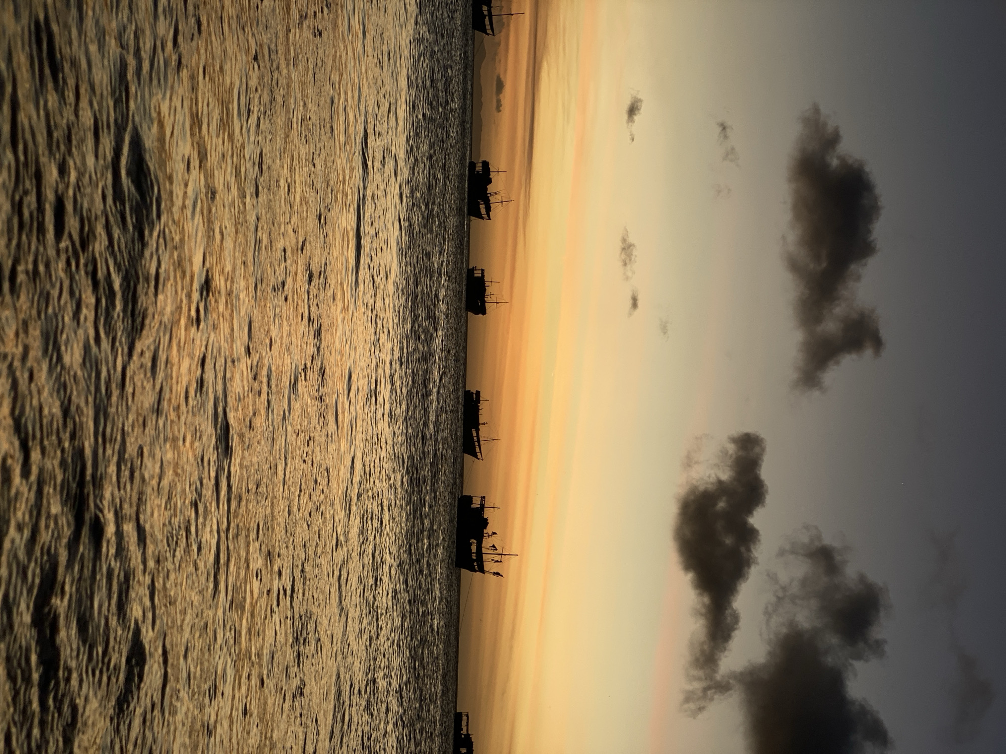The image depicts a seascape in Juan Griego, Venezuela, during a sunset or sunrise, as evidenced by the warm, golden hues in the sky and reflected on the water's surface. Several silhouetted boats are scattered along the horizon, suggesting a fishing or maritime activity. The sky is partially covered by dark, billowy clouds. The sea has a rippled texture, illuminated by the sunlight. The overall composition suggests a tranquil, coastal scene.